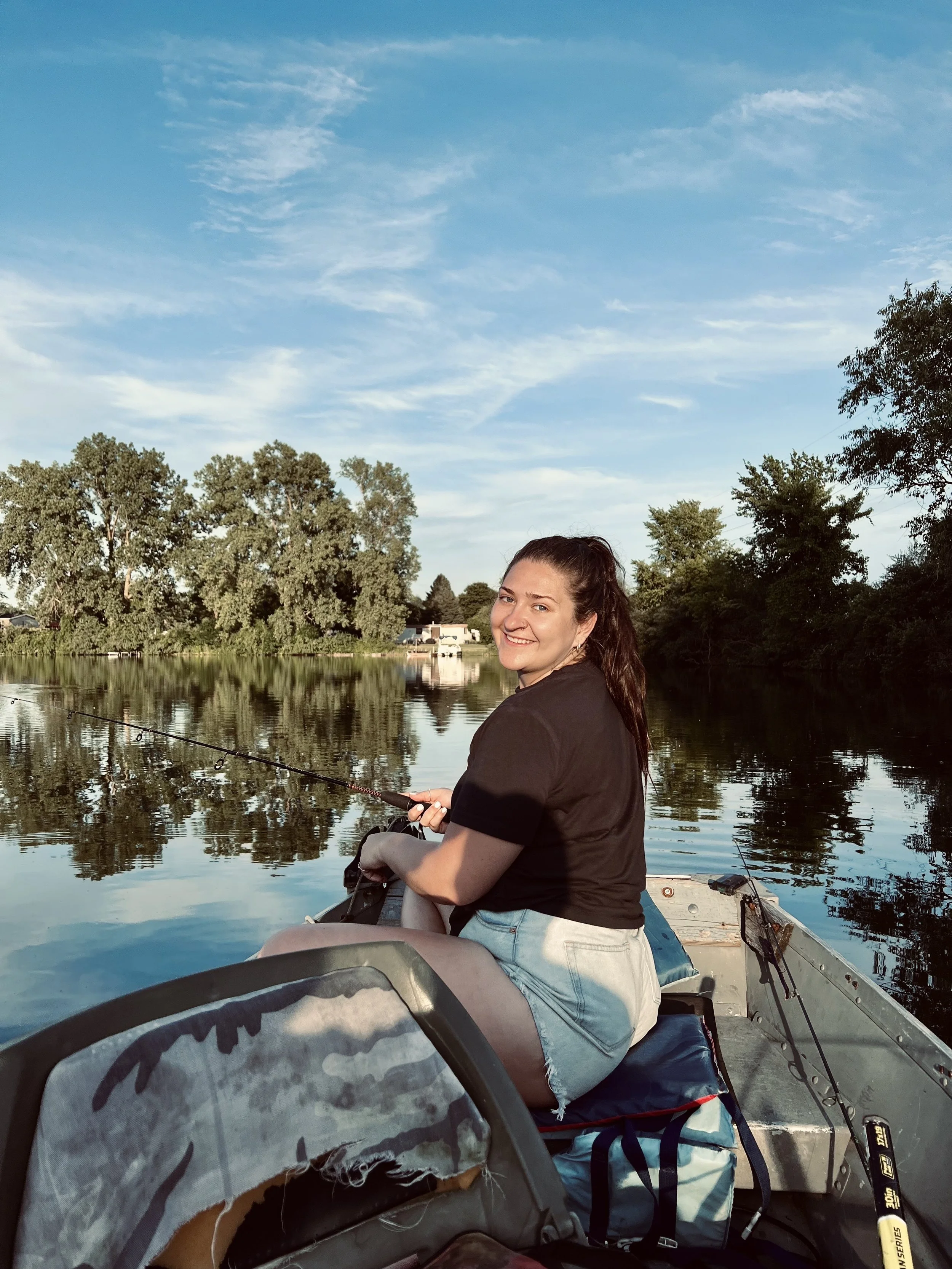 A woman sitting in a boat fishing on a calm river during sunset, surrounded by trees and blue sky.