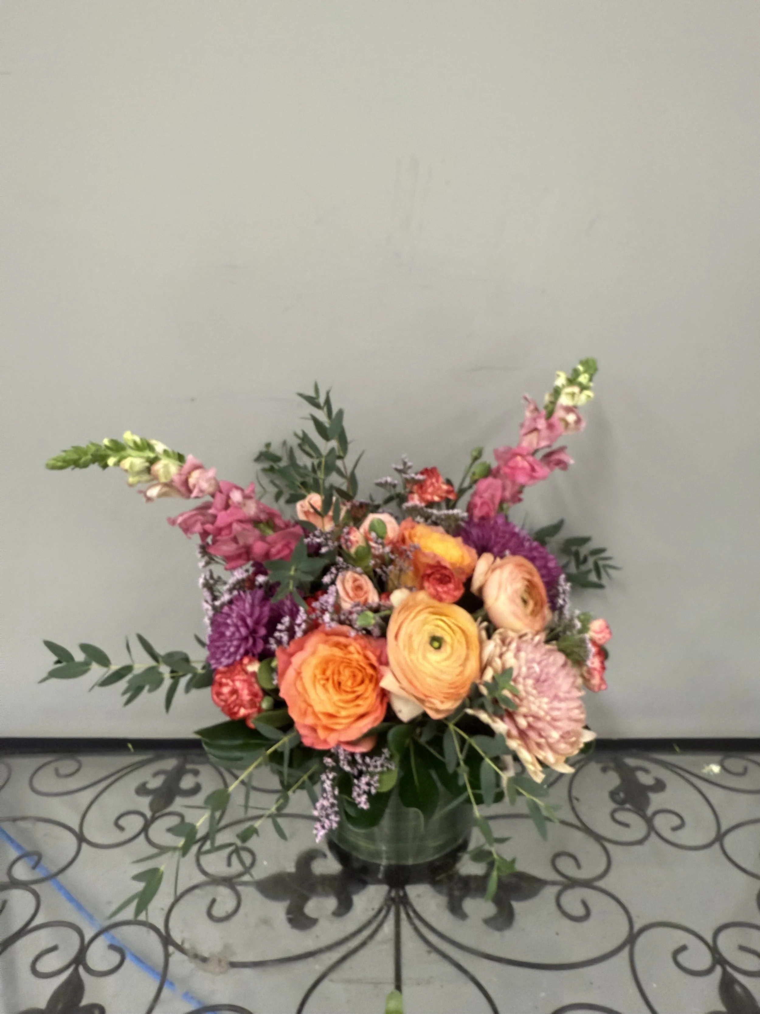 A colorful bouquet of flowers in a glass vase, including roses, ranunculus, and other blooms with greenery, placed on a decorative metal table.