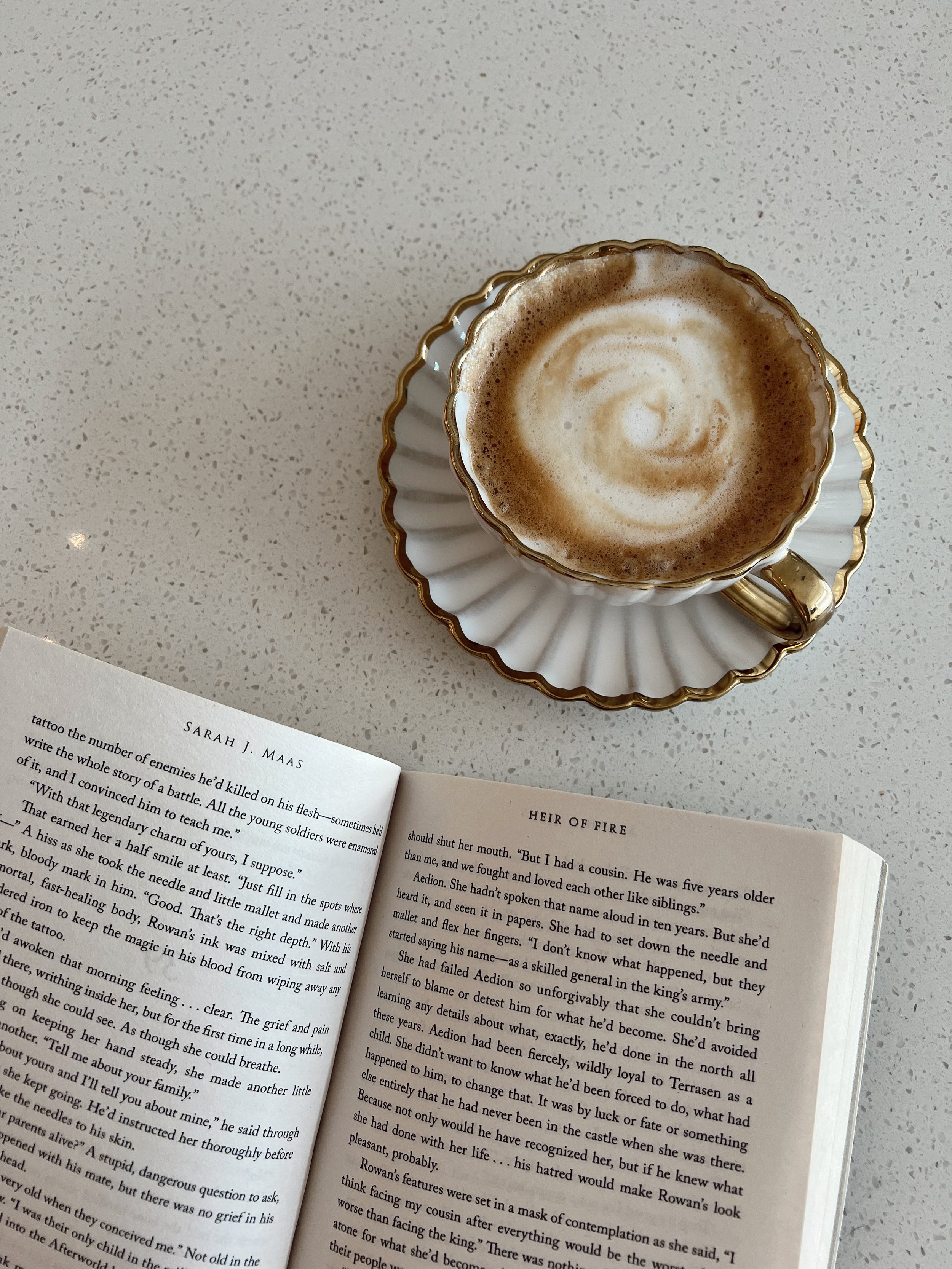 A cup of coffee with frothy milk in a decorative saucer, placed on a speckled white countertop, next to an open book.