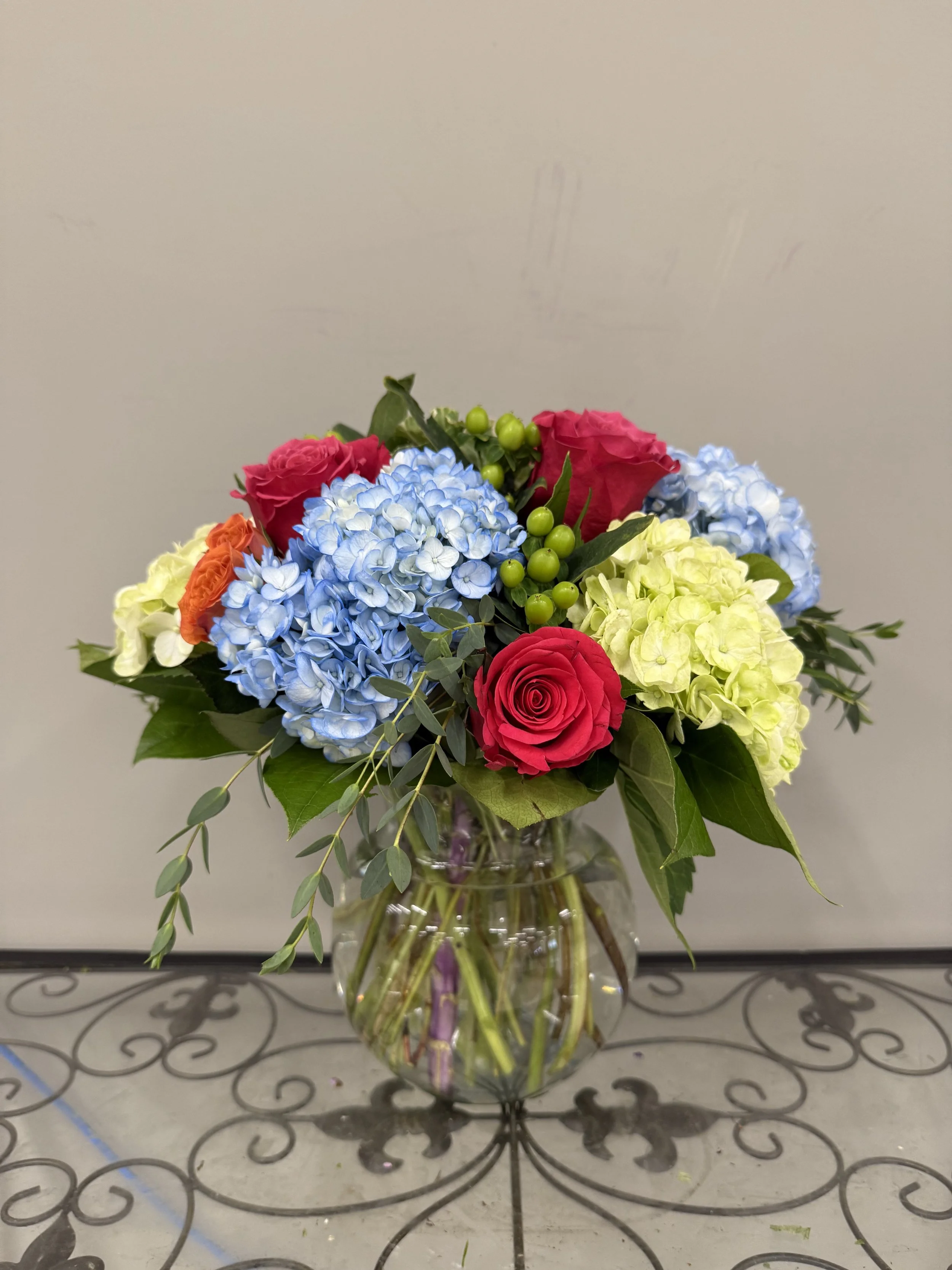 A colorful bouquet of flowers in a clear glass vase, including pink roses, light blue hydrangeas, yellow hydrangeas, orange flowers, green hypericum berries, and assorted greenery, placed on a decorative metal table.