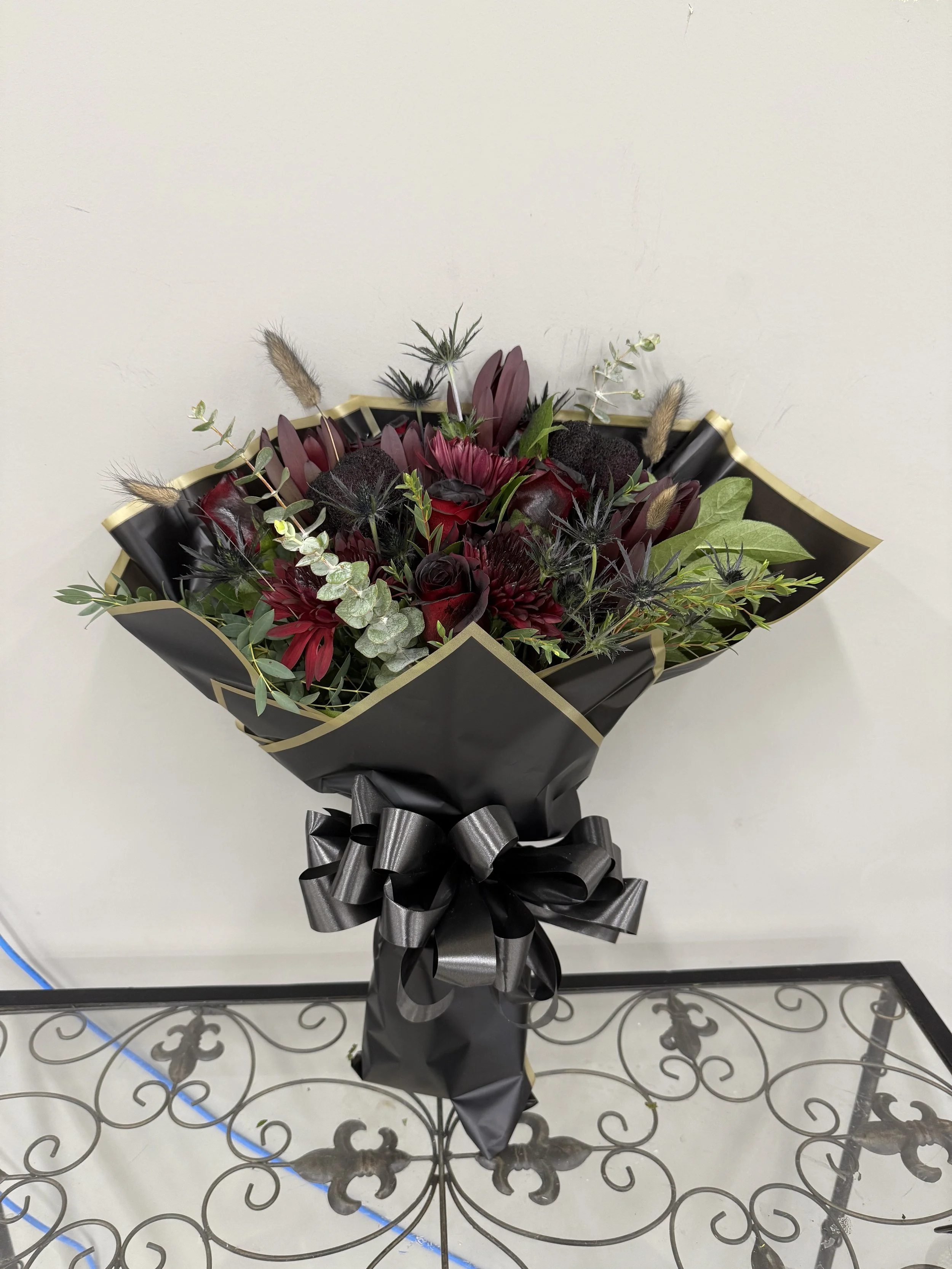 A bouquet of dark red and black flowers with green foliage, wrapped in black paper with a gold edge, and tied with a black ribbon, placed on a decorative table.