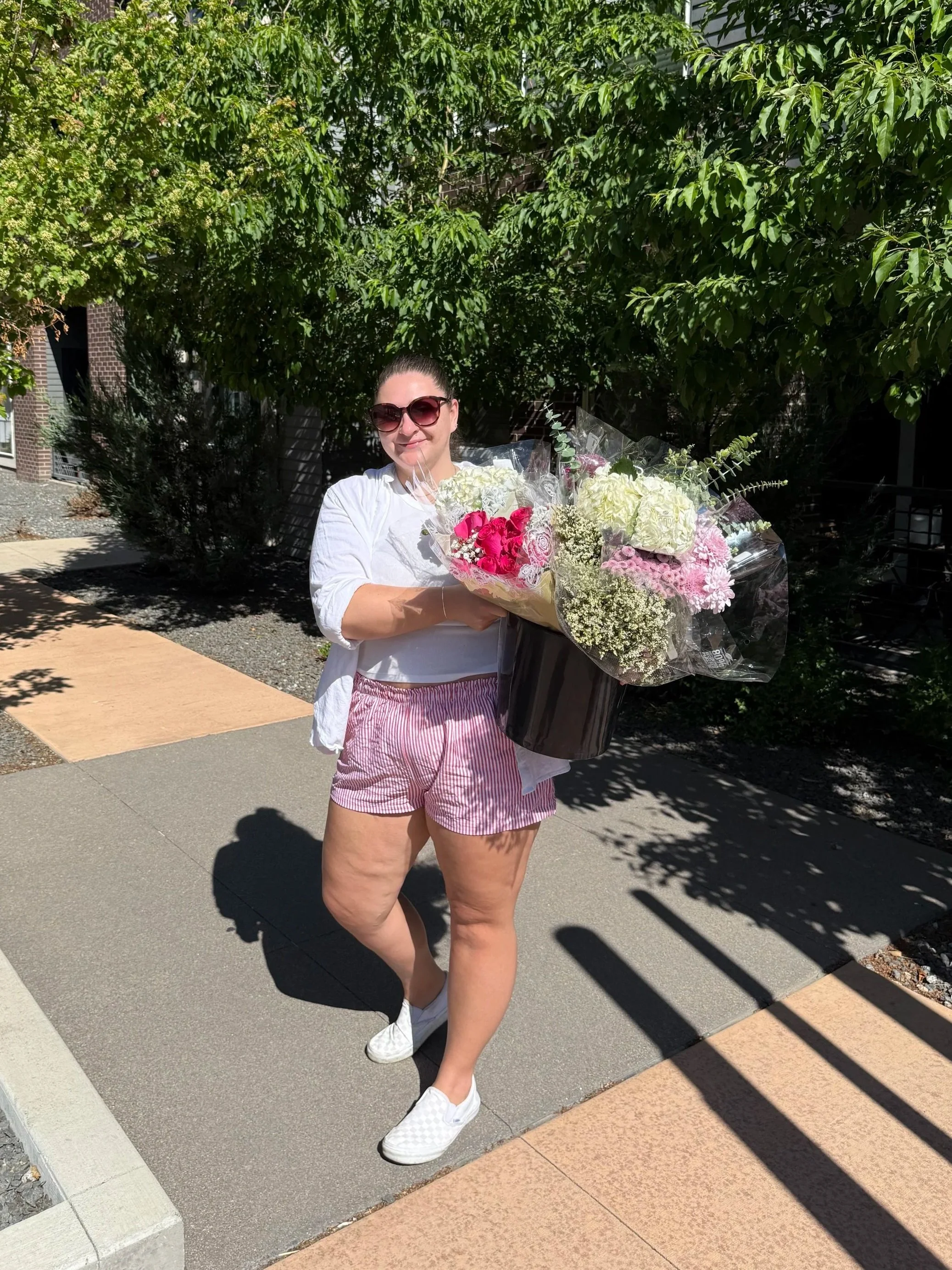 A woman wearing sunglasses, a white shirt, pink and white striped shorts, and white shoes is standing outdoors on a sidewalk, holding a large bouquet of pink and white flowers wrapped in clear plastic. She is smiling and standing in front of green leafy trees.