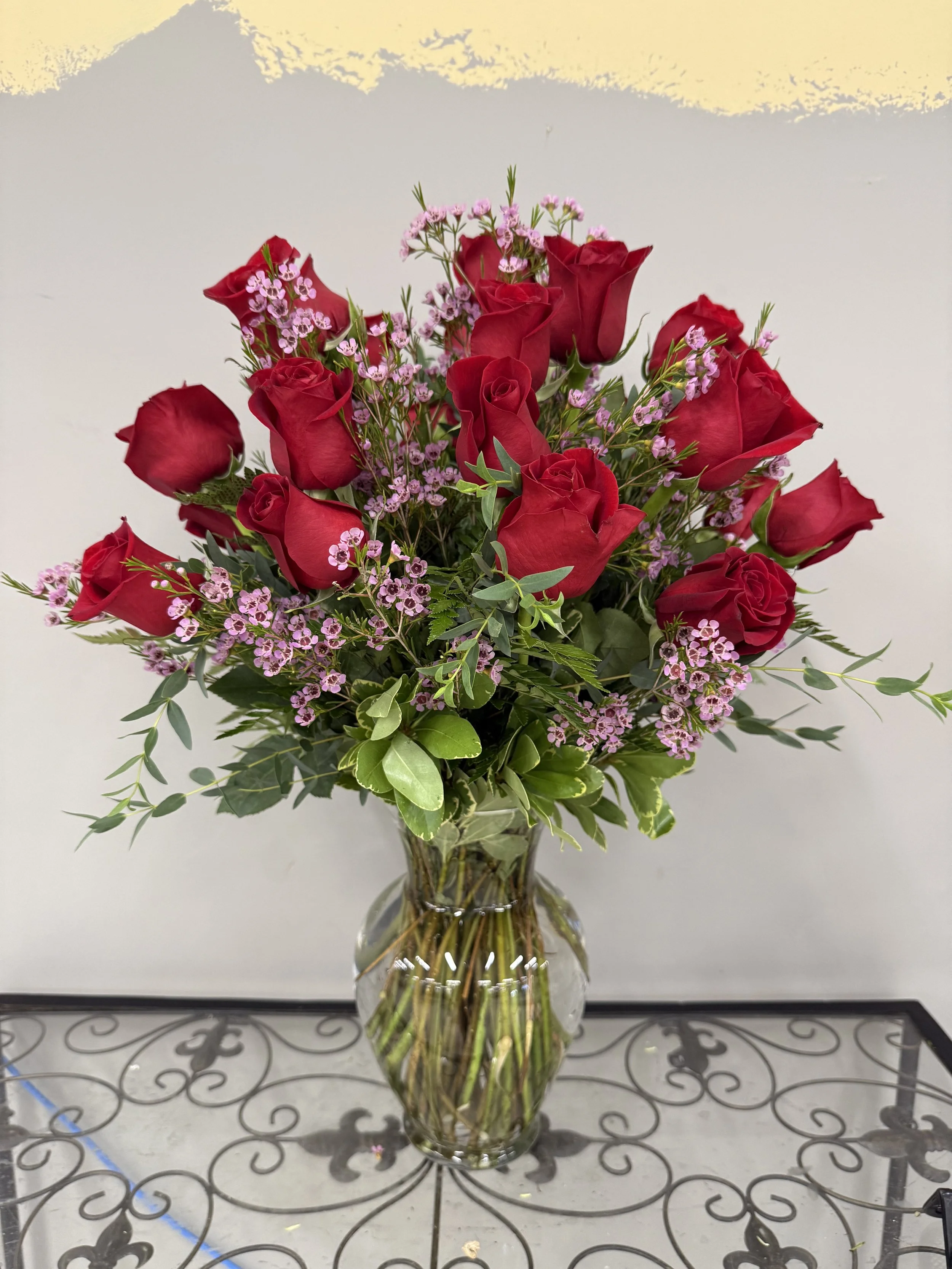 A clear glass vase filled with red roses, pink small flowers, and green foliage on a decorative metal table against a plain light-colored wall.