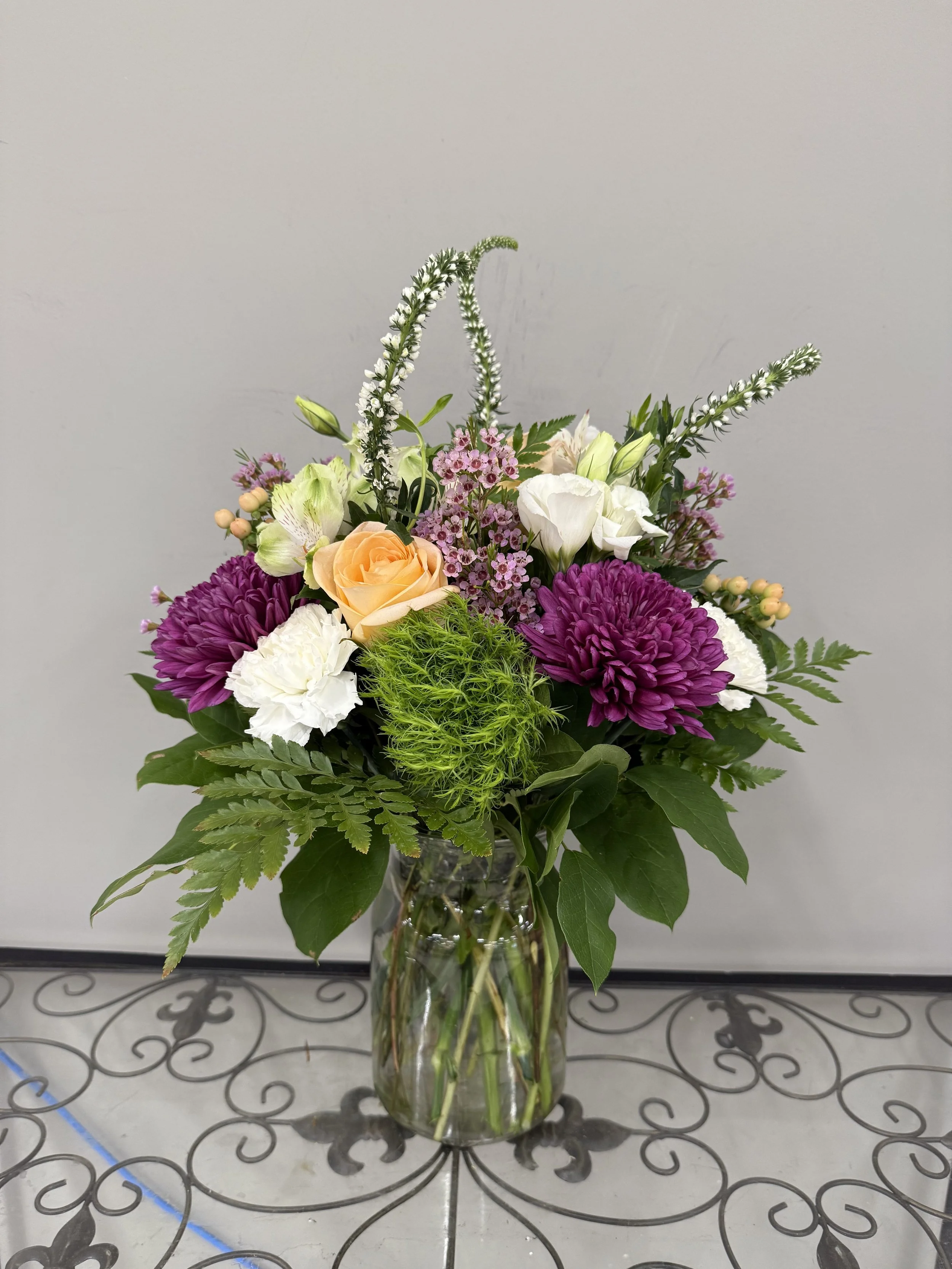 A colorful flower bouquet in a clear glass vase containing roses, dahlias, lilacs, ferns, and other assorted flowers on a decorative metal table.