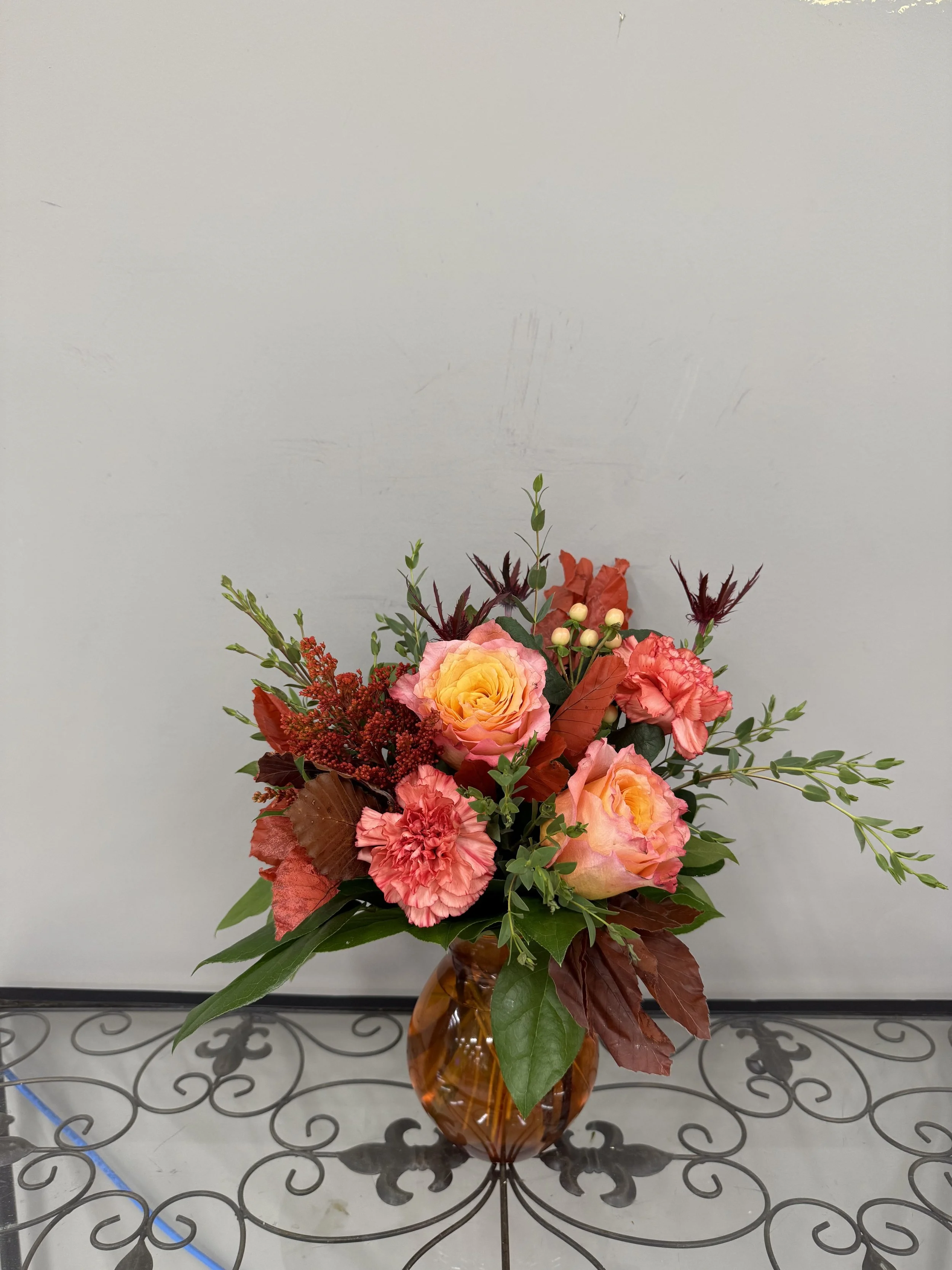 A floral arrangement with pink roses, red carnations, and various green and reddish foliage in a brown glass vase placed on a decorative metal table against a plain light-colored wall.