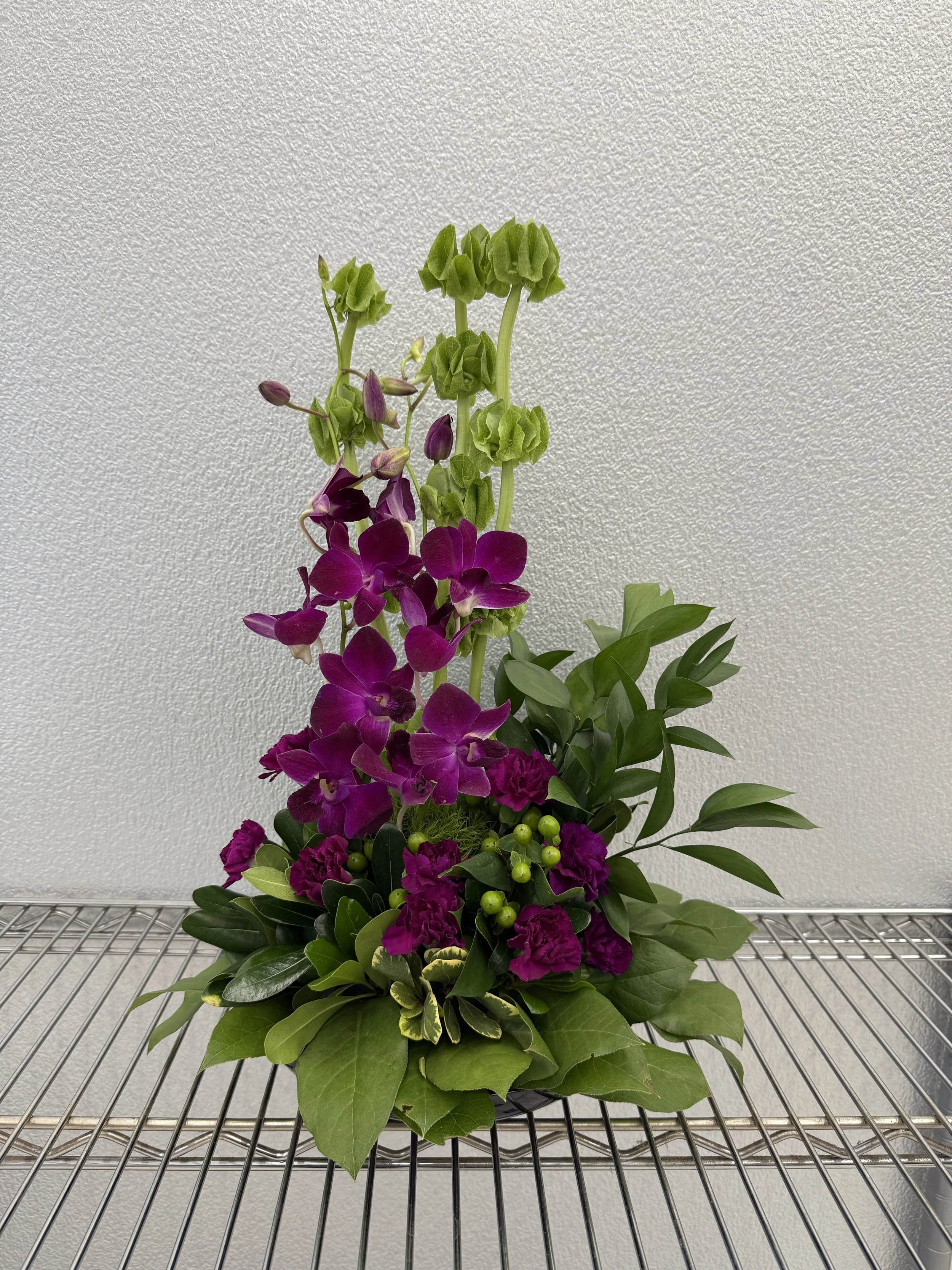 A floral arrangement with purple orchids, pink carnations, green foliage, and berries, placed on a metal wire shelf against a plain white wall.