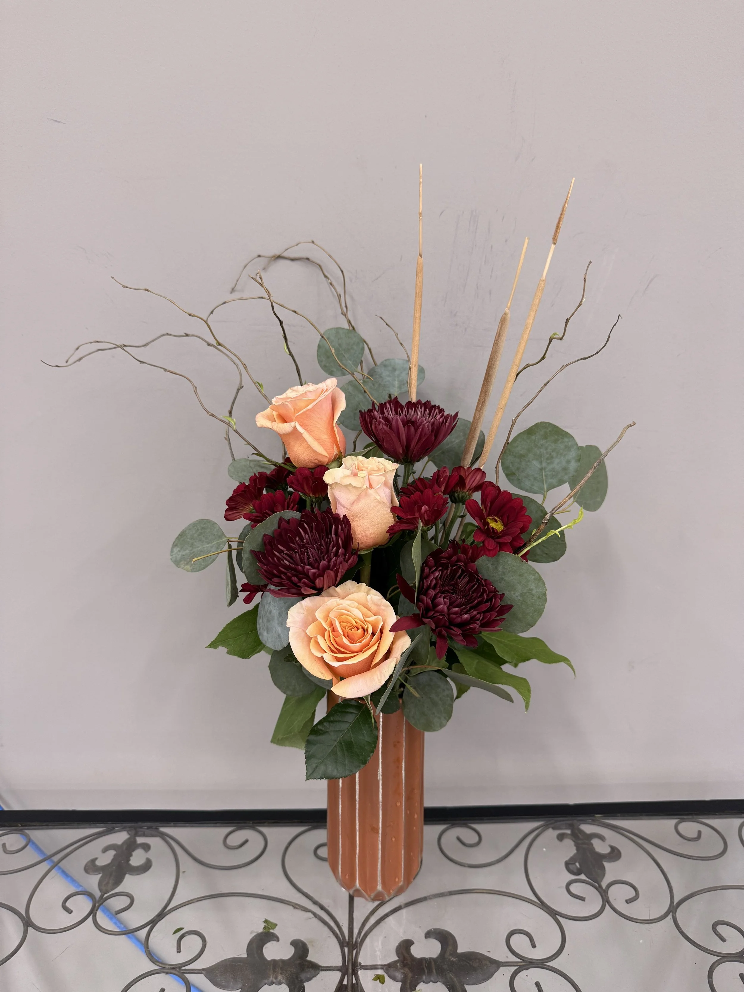 A floral arrangement with peach roses, dark red chrysanthemums, eucalyptus leaves, and curly twigs in a tall, ridged, terracotta-colored vase, set on a decorative metal table against a gray wall.
