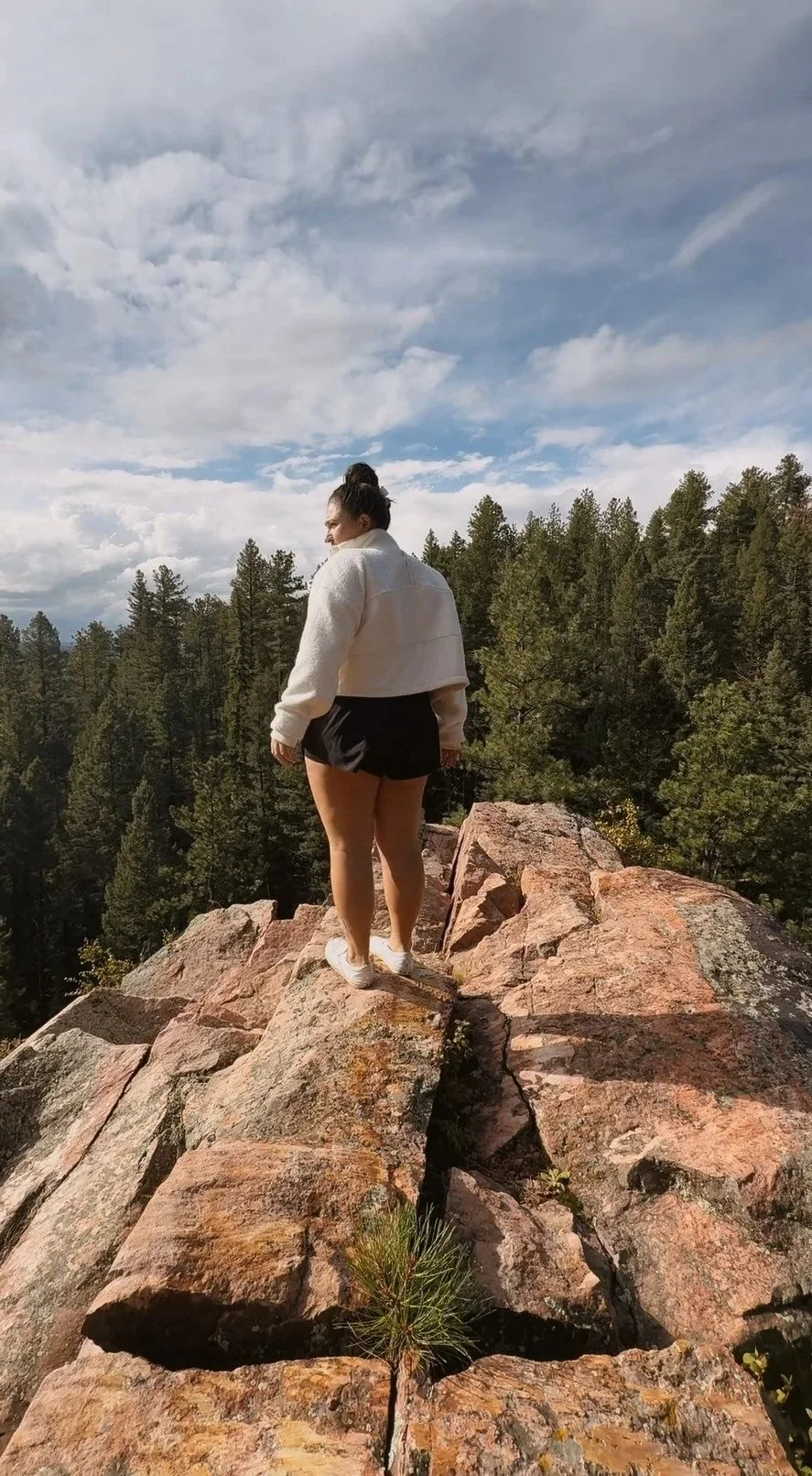 A woman in a cream jacket and black shorts walking on rocks on a mountain with trees and a cloudy sky in the background.