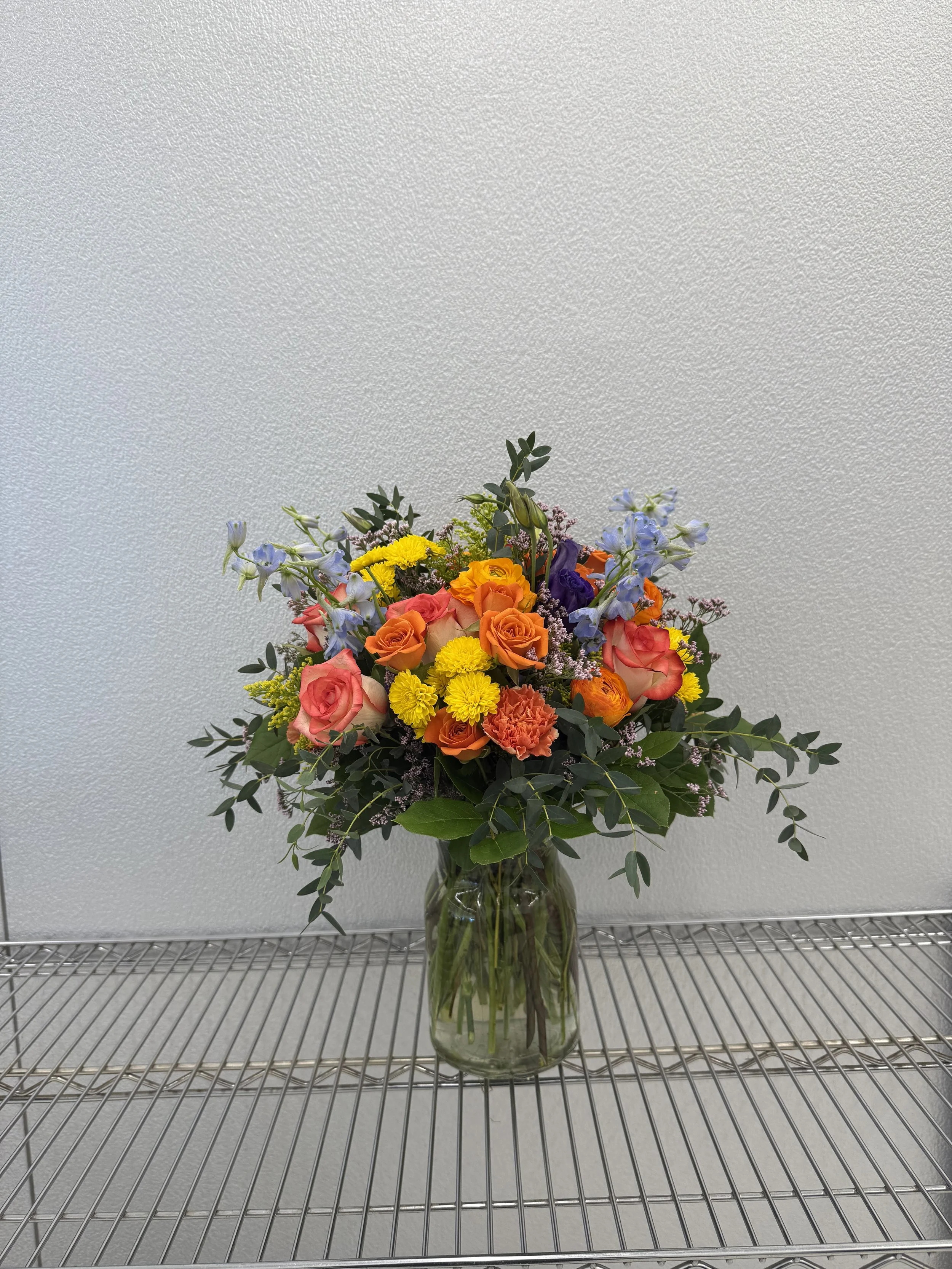 A colorful bouquet of flowers in a glass vase on a metal shelf against a plain textured wall.
