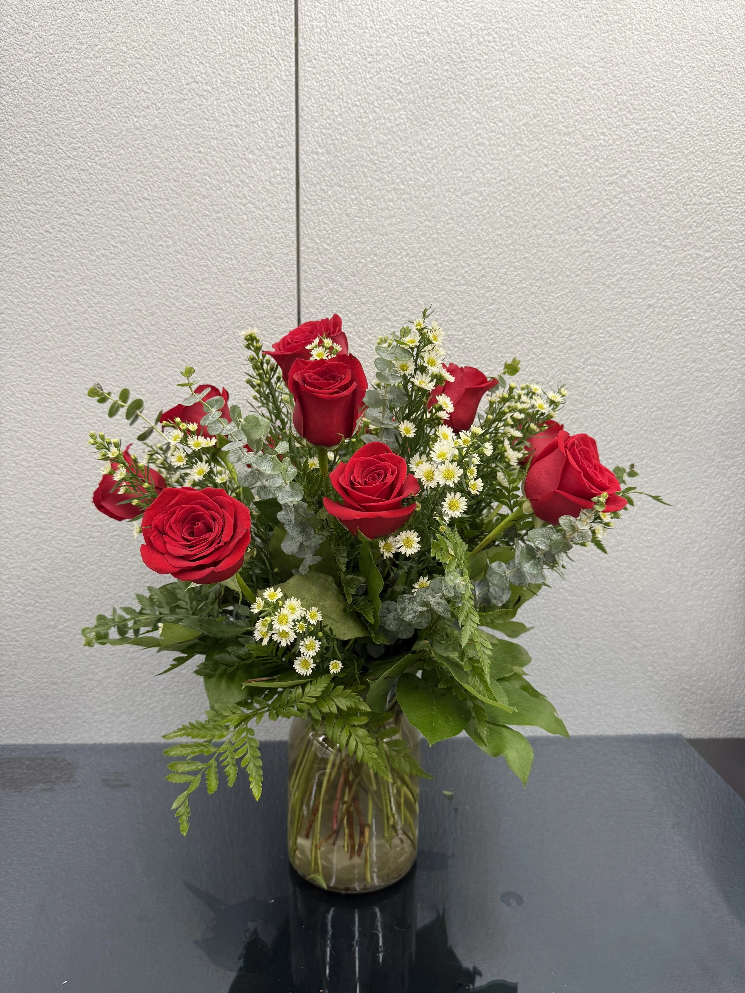A bouquet of red roses, small white daisies, and green foliage in a glass vase on a dark table against a plain white wall.