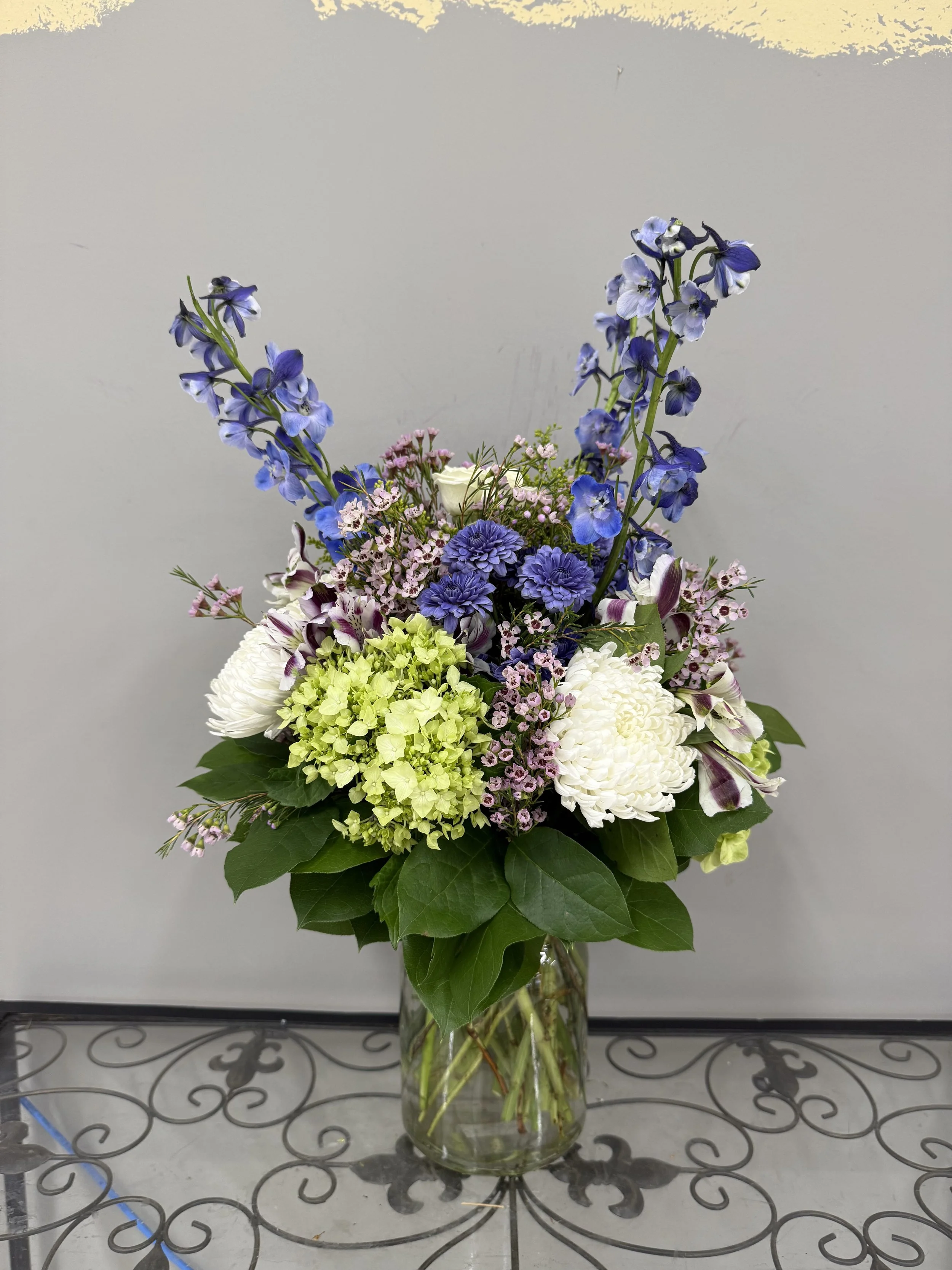 A floral arrangement in a glass vase with purple, white, and green flowers and green leaves.