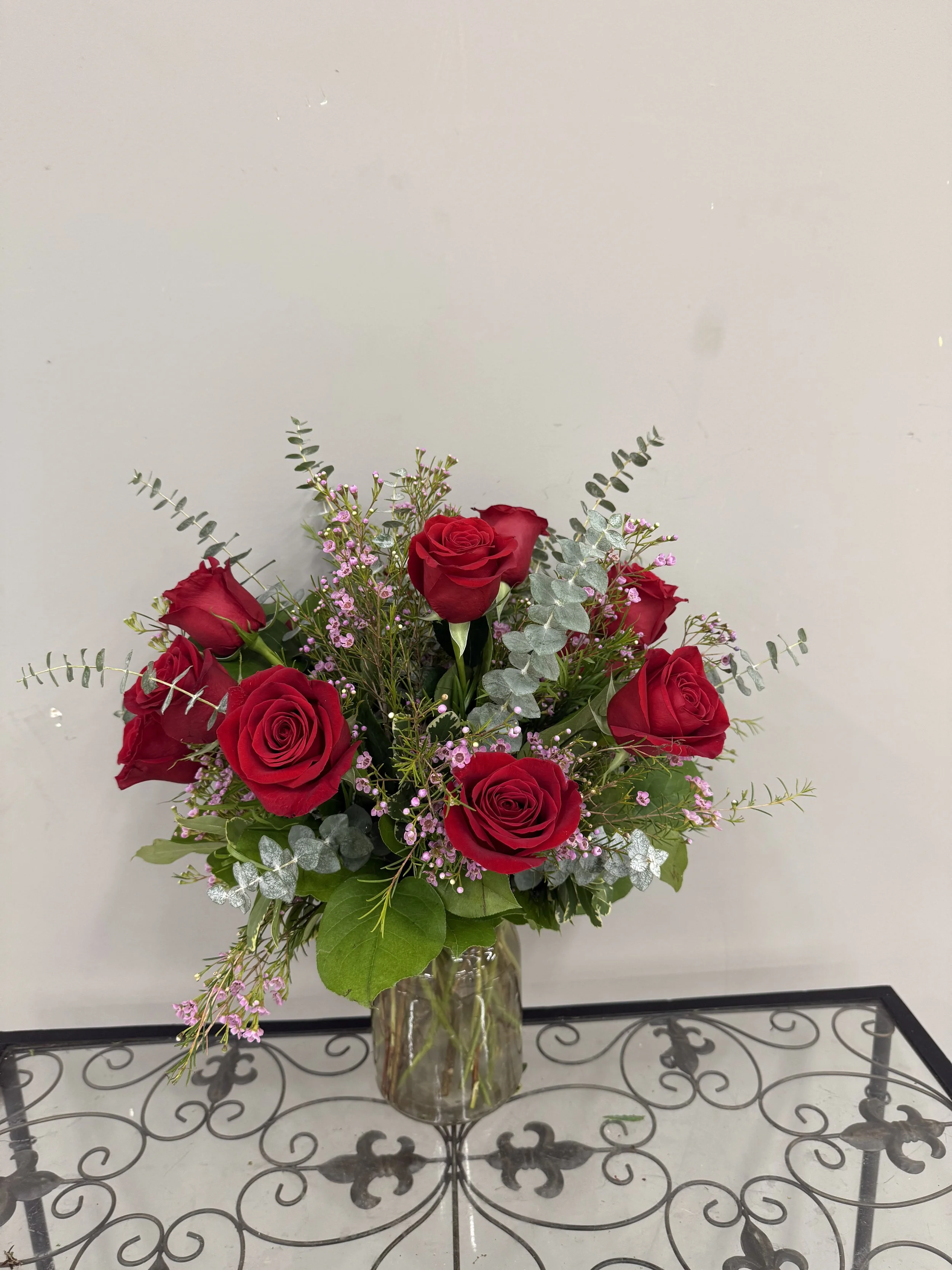 A bouquet of red roses with pink filler flowers and greenery in a glass vase on a metal table against a plain light-colored wall.