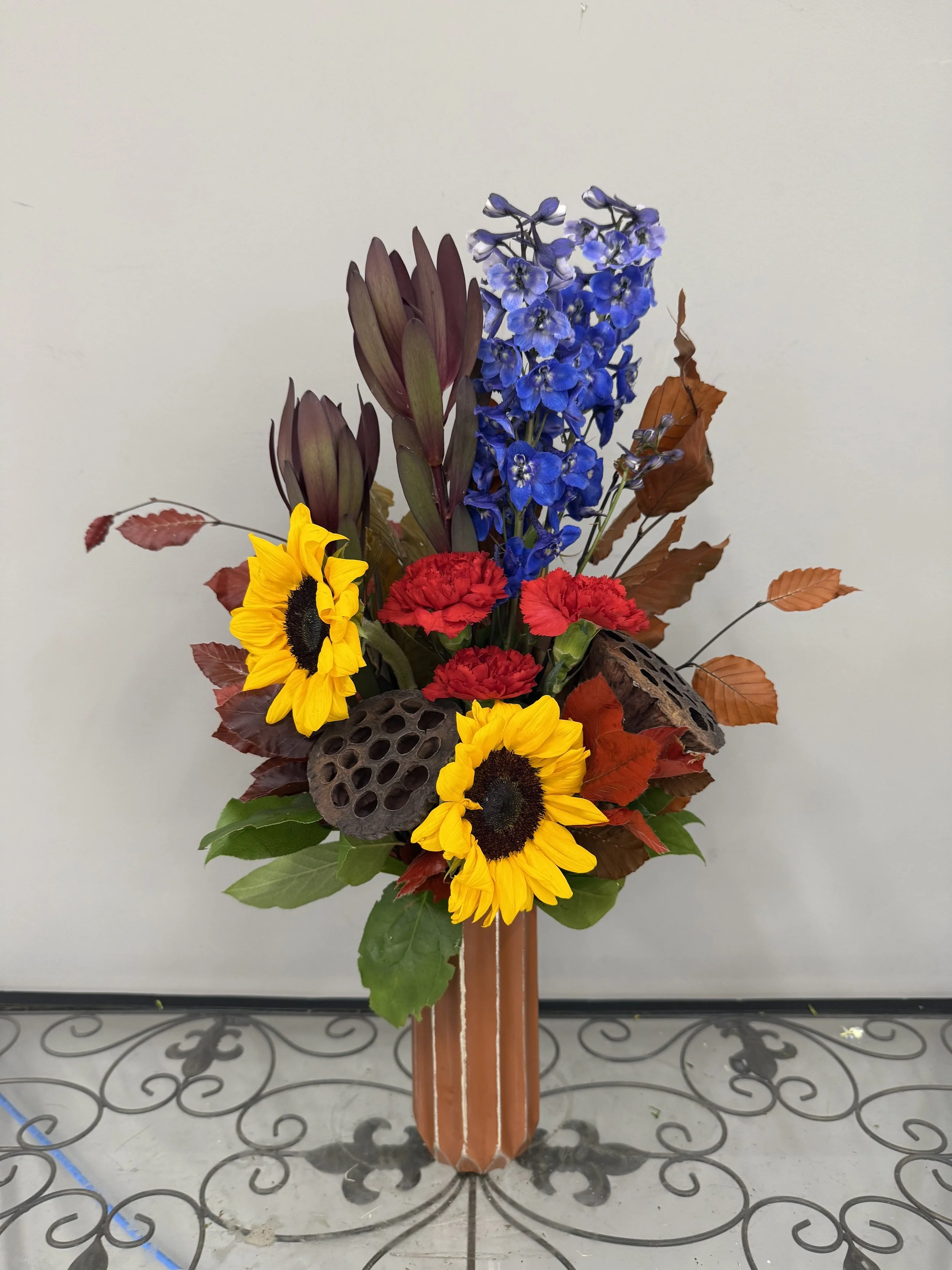A flower arrangement in a brown striped vase with sunflowers, red carnations, blue delphiniums, purple-tinted leaves, and lotus pods, set on a decorative black metal table against a plain white wall.