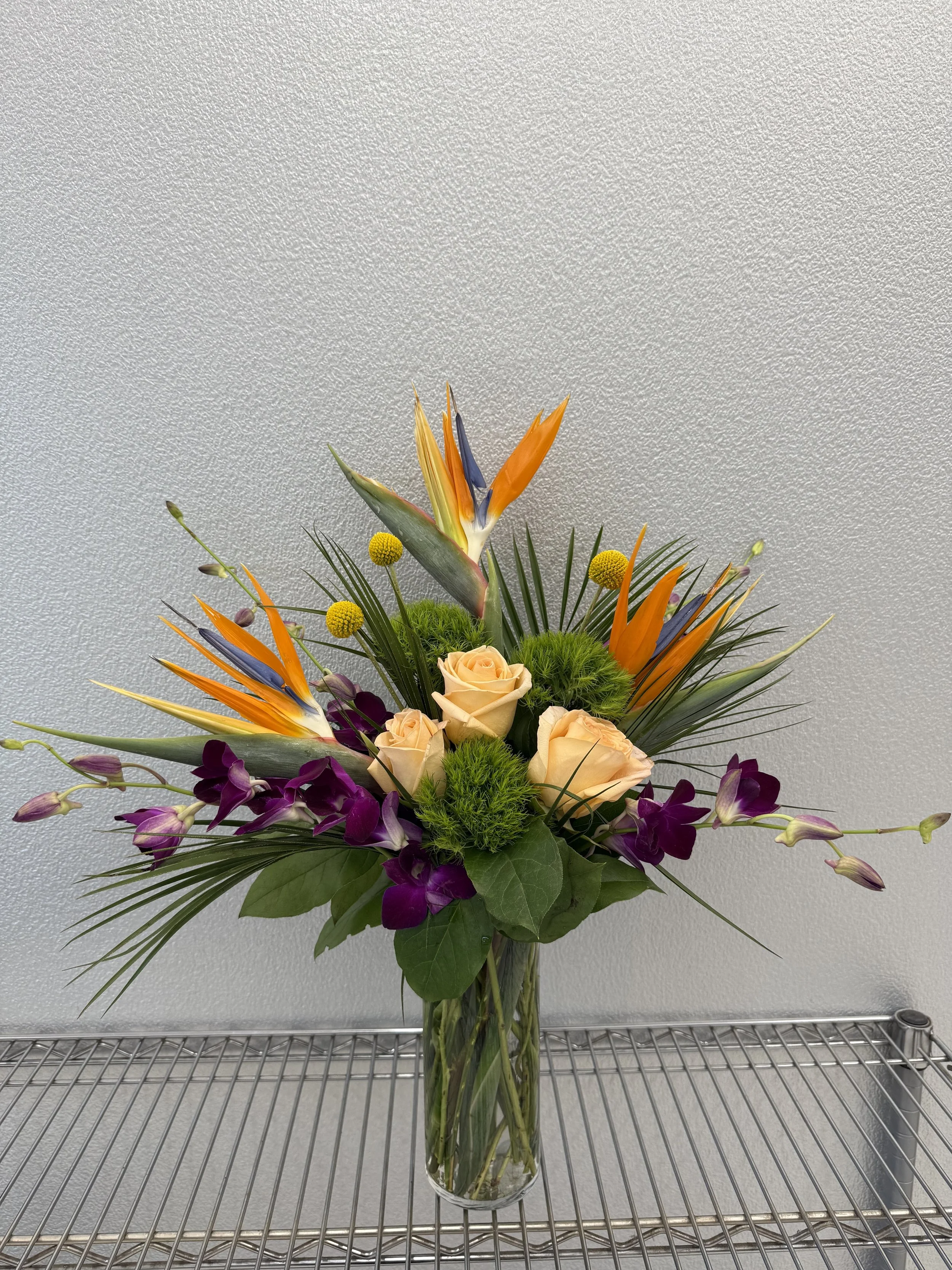 A colorful flower arrangement in a tall glass vase, featuring peach roses, purple orchids, orange bird of paradise flowers, green foliage, and small yellow round flowers against a plain light gray background.