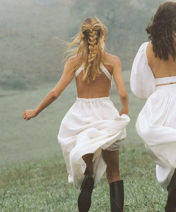 Two women running outdoors in a grassy field, wearing white dresses. One woman has long blonde hair with a braid, and the other woman has shoulder-length brown hair.