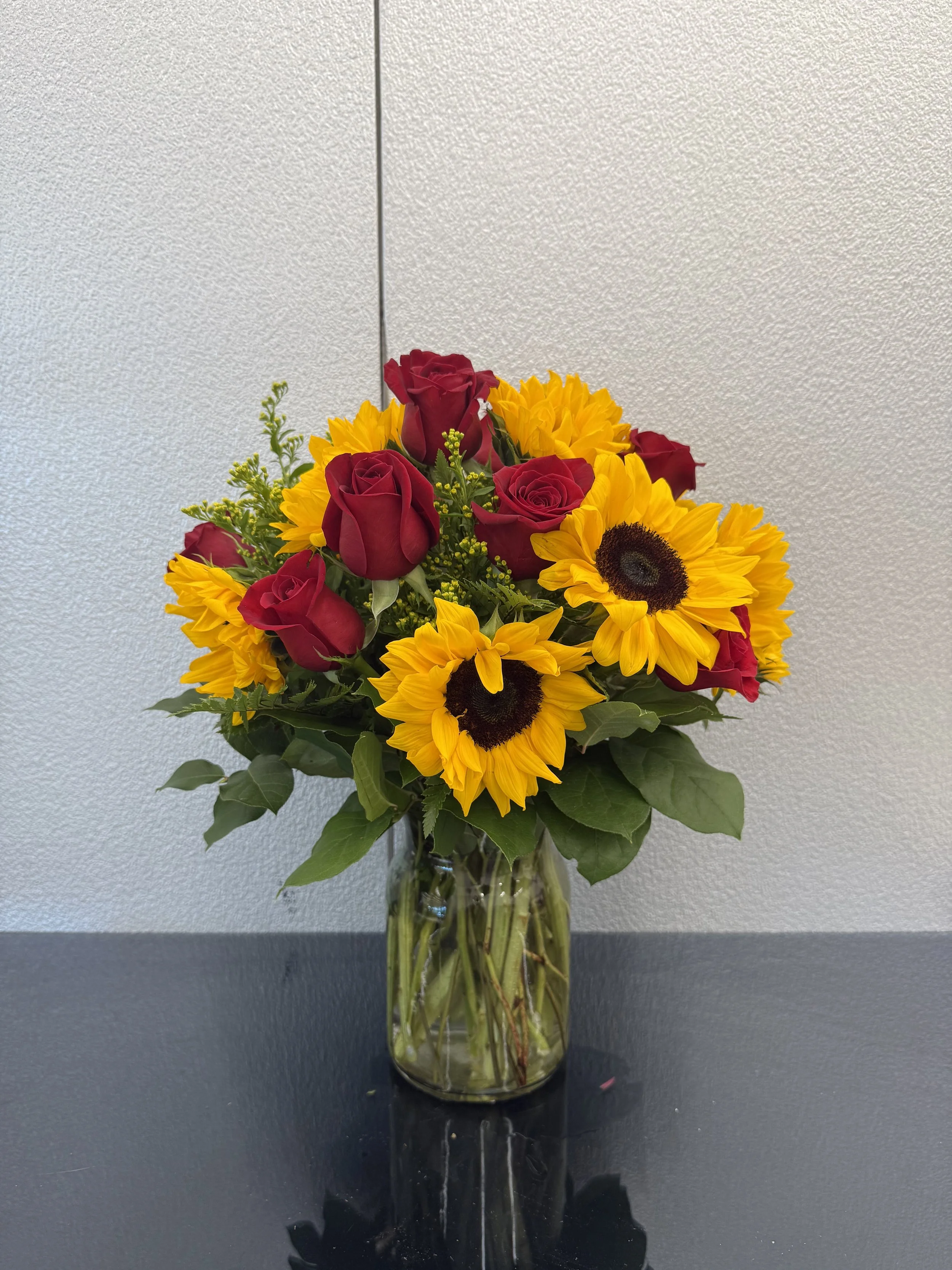 Vase of red roses, yellow sunflowers, and green foliage on a black surface against a textured light gray wall.