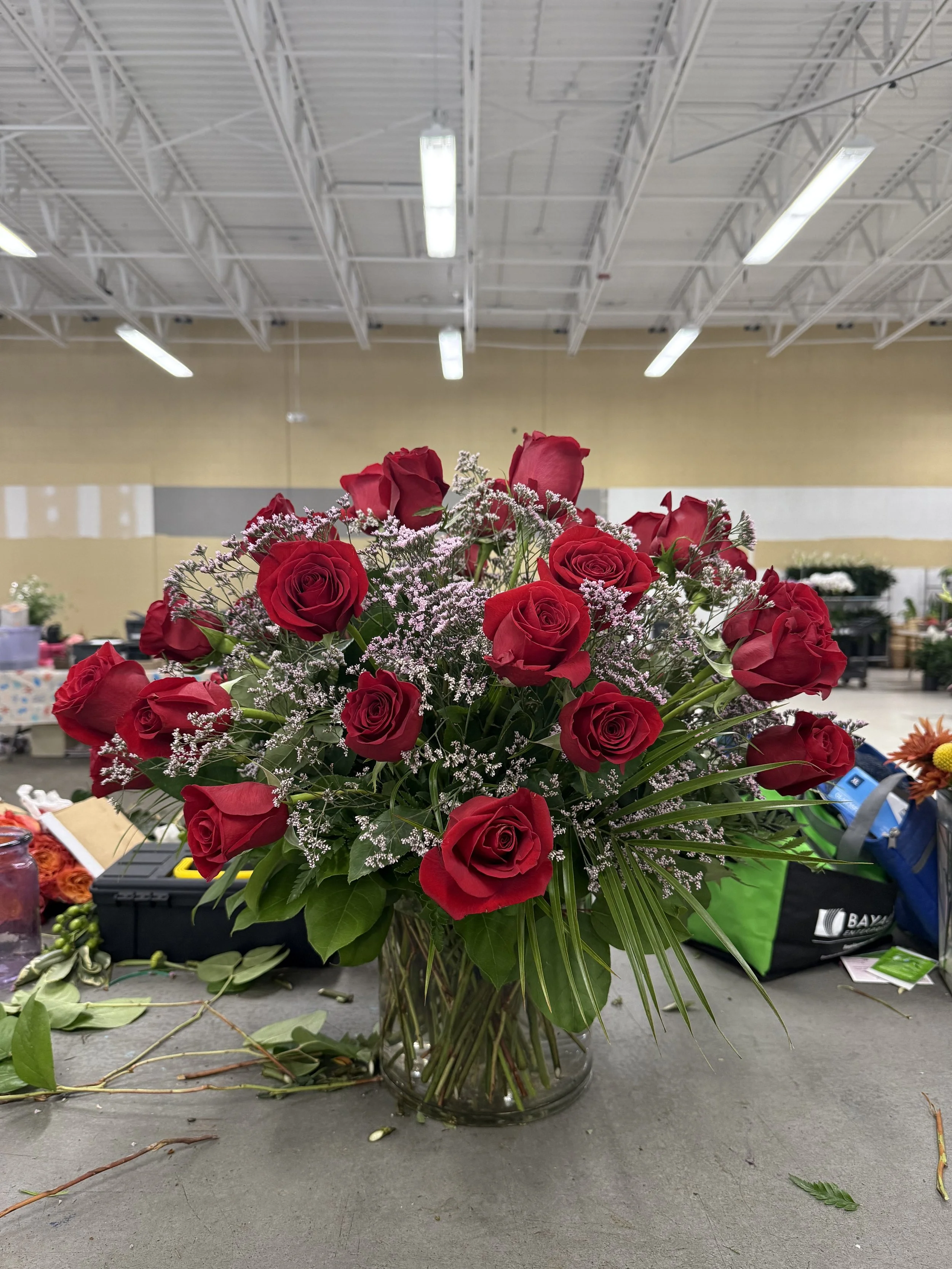 A bouquet of red roses with white filler flowers in a glass vase on a table, with a flower market in the background.