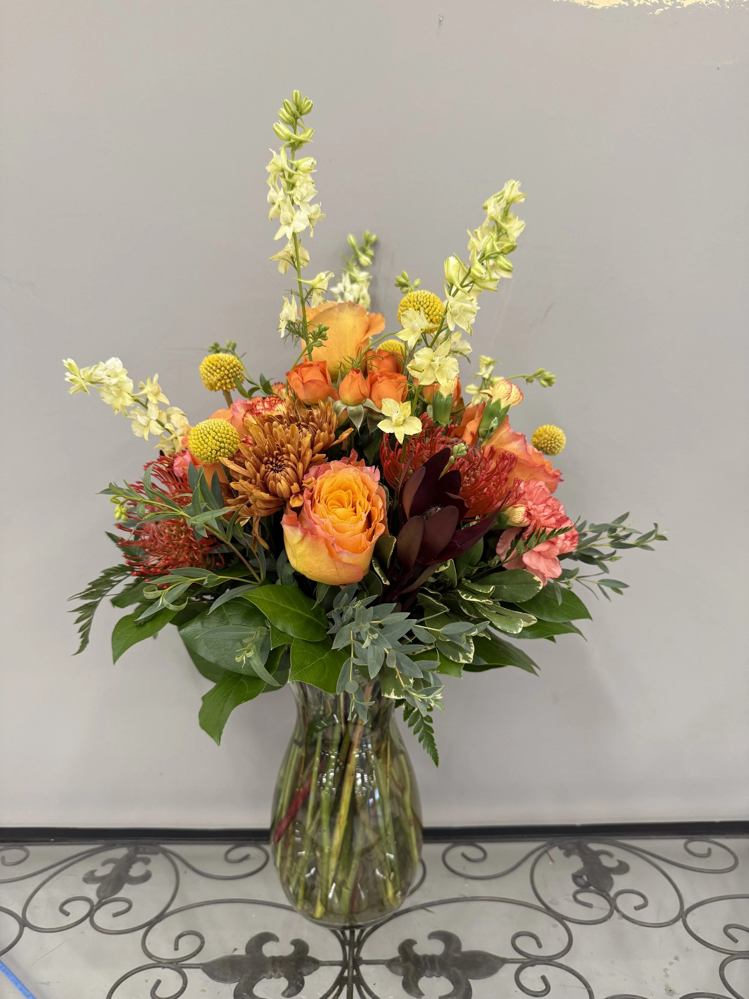A colorful bouquet of flowers in a clear glass vase on a metal table, featuring roses, lilies, chrysanthemums, and various greenery.