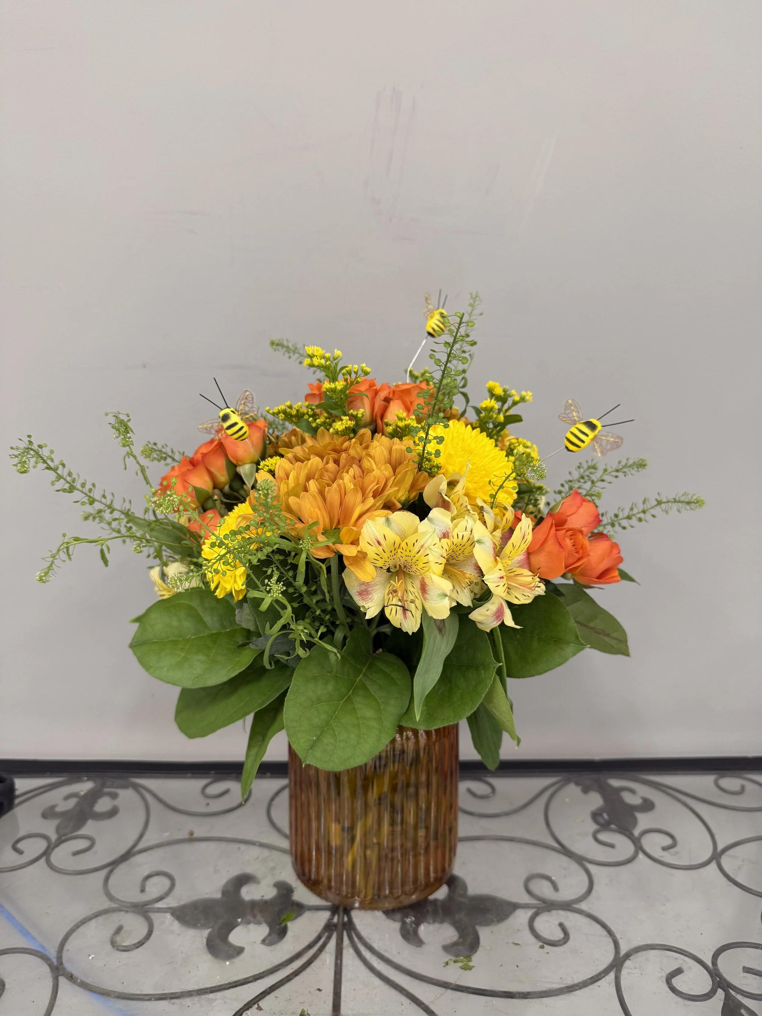 A colorful flower arrangement in a brown glass vase, featuring yellow, orange, and pale yellow flowers, accented with green leaves and small bees.