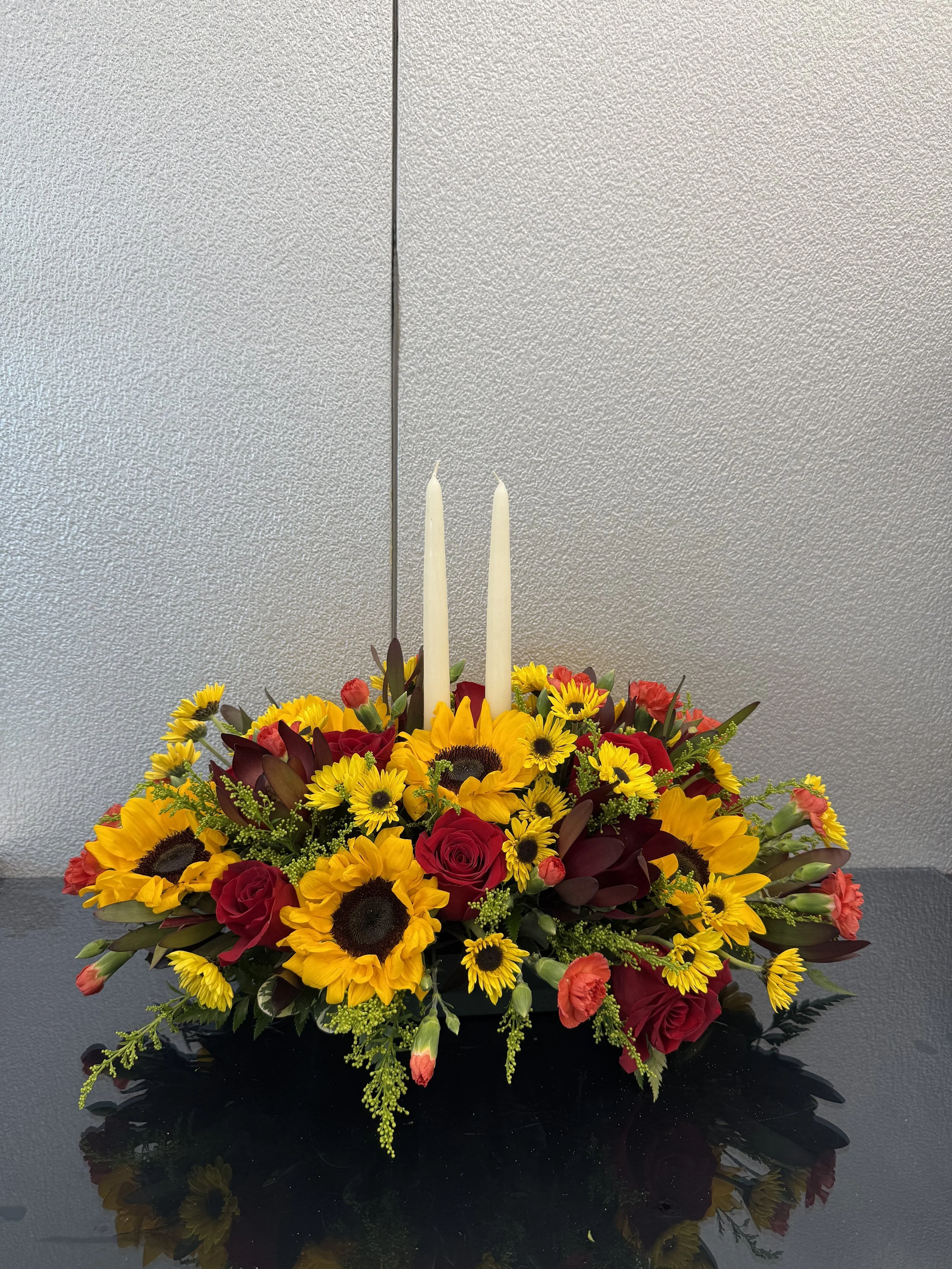 A floral arrangement with yellow sunflowers, red roses, and yellow daisies, topped with two white candles, placed on a black reflective surface against a gray textured background.