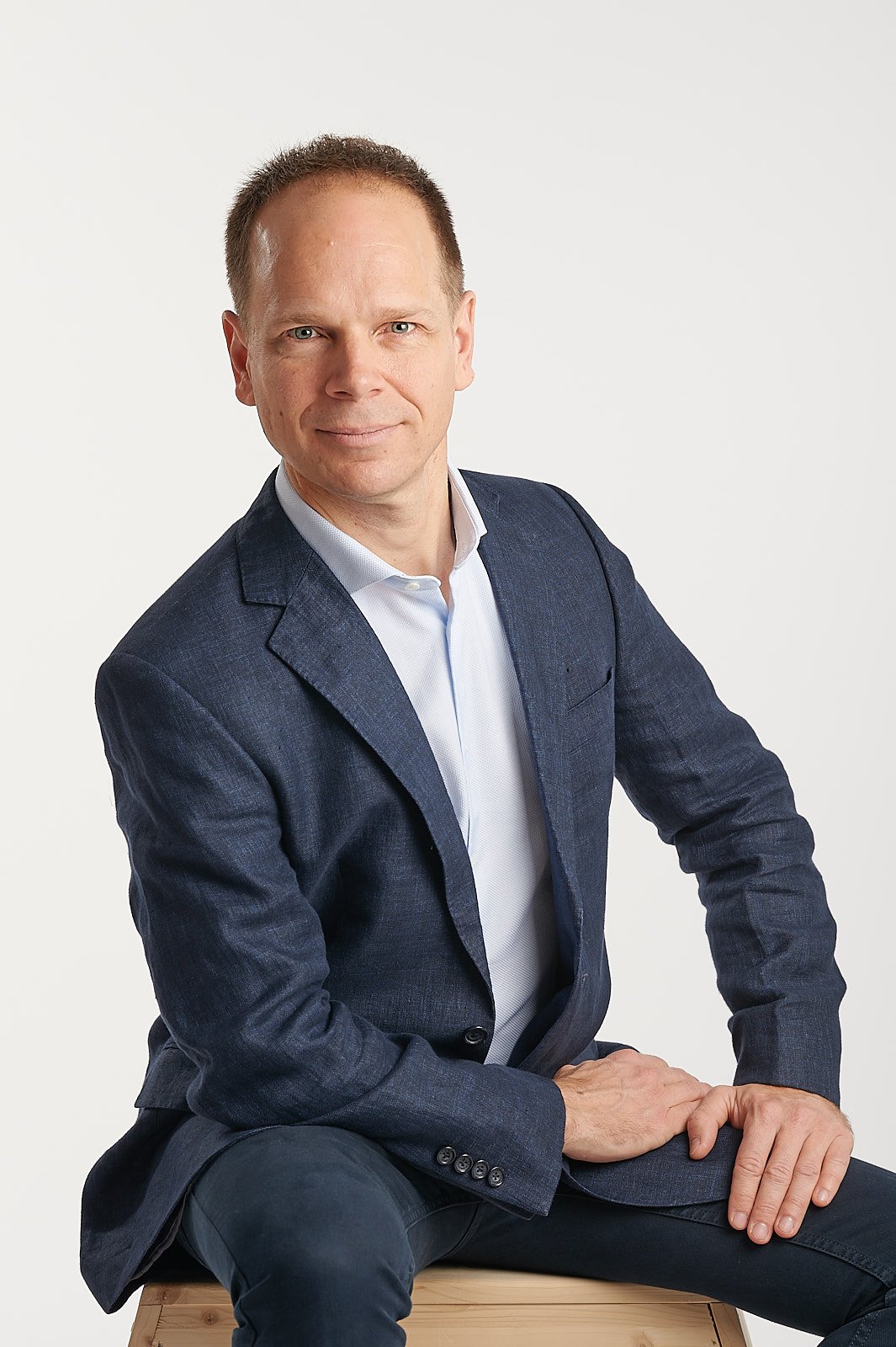 A man with short hair wearing a blue blazer and white shirt, sitting on a wooden surface, posing for a professional portrait against a plain white background.