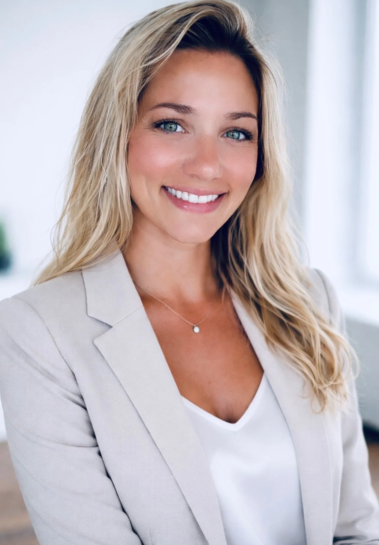 A woman with long blonde hair smiling, wearing a beige blazer and white top, in a bright indoor setting.