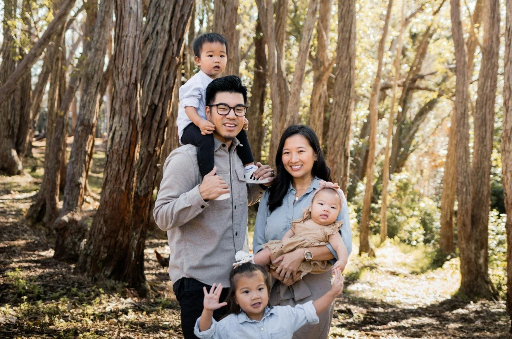 A family of five posing outdoors in a wooded area with tall trees, including a father, mother, two young children, and a toddler sitting on the father's shoulders.