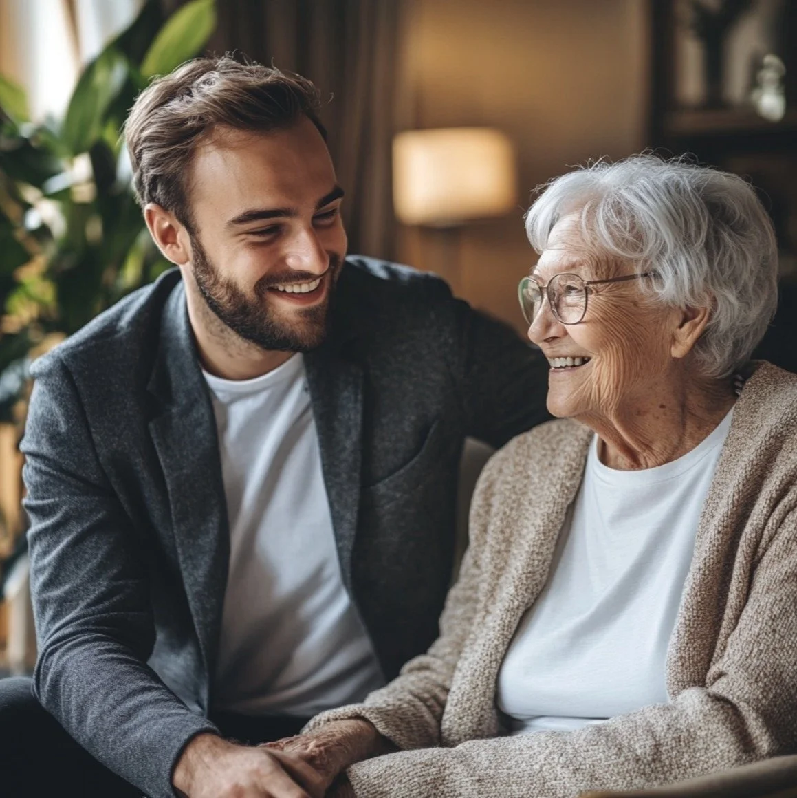 A young man and an elderly woman share a happy moment sitting together indoors. The young man is smiling at the elderly woman, and she is smiling back. They appear to be enjoying each other's company, with a warm and cozy background that includes a lamp and plants.