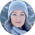 Close-up photo of a woman with gray hair wearing a blue knit hat and scarf, outdoors in a winter setting with snow-covered trees in the background.