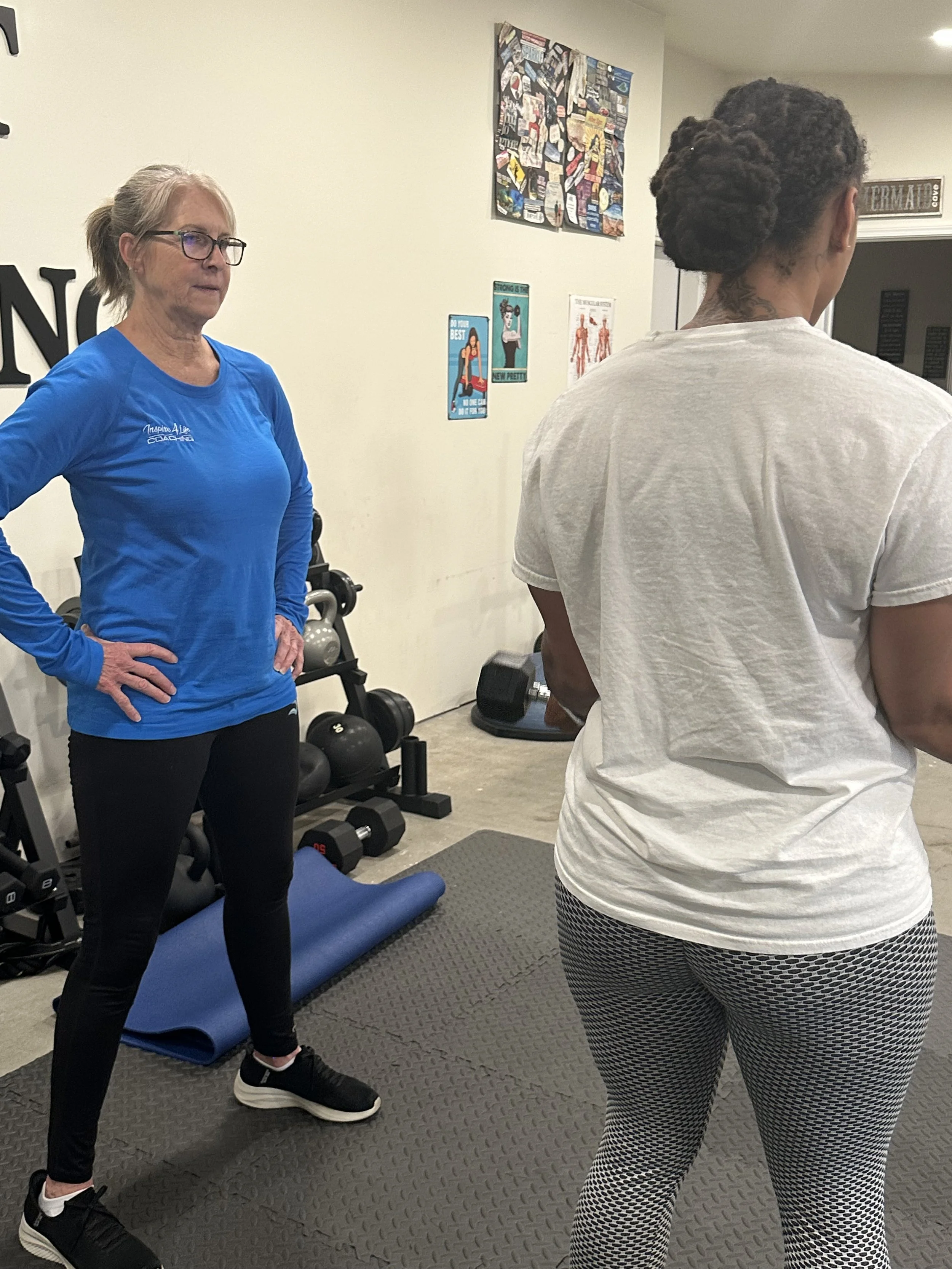 A woman wearing a blue long-sleeve shirt and black leggings standing in a gym, facing another woman in a white t-shirt and patterned leggings. The gym has workout posters on the wall and various exercise equipment.