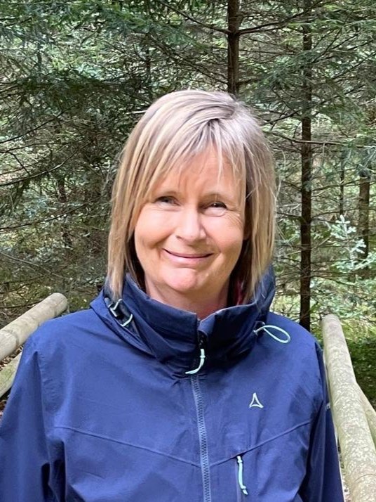 Frau in blauer Outdoor-Jacke im Wald auf einem Holzweg, umgeben von Bäumen.