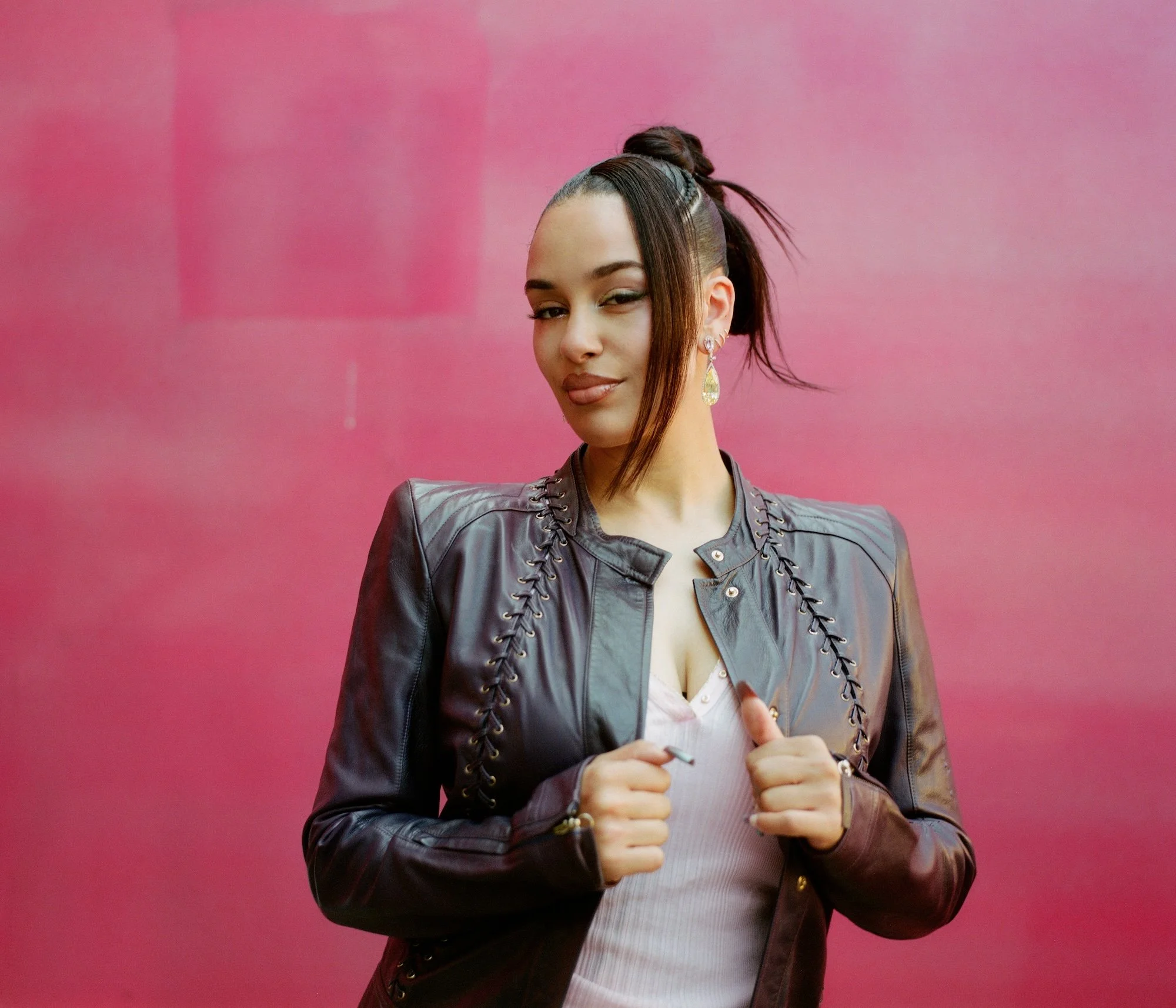 Young woman with a high ponytail, wearing a leather jacket and casual clothing, standing against a pink background, giving a thumbs-up gesture with both hands.