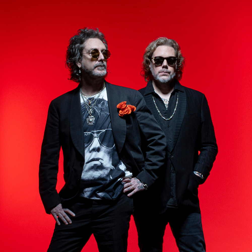 Two men posing; one wearing a patterned jacket and sunglasses, the other holding a guitar.