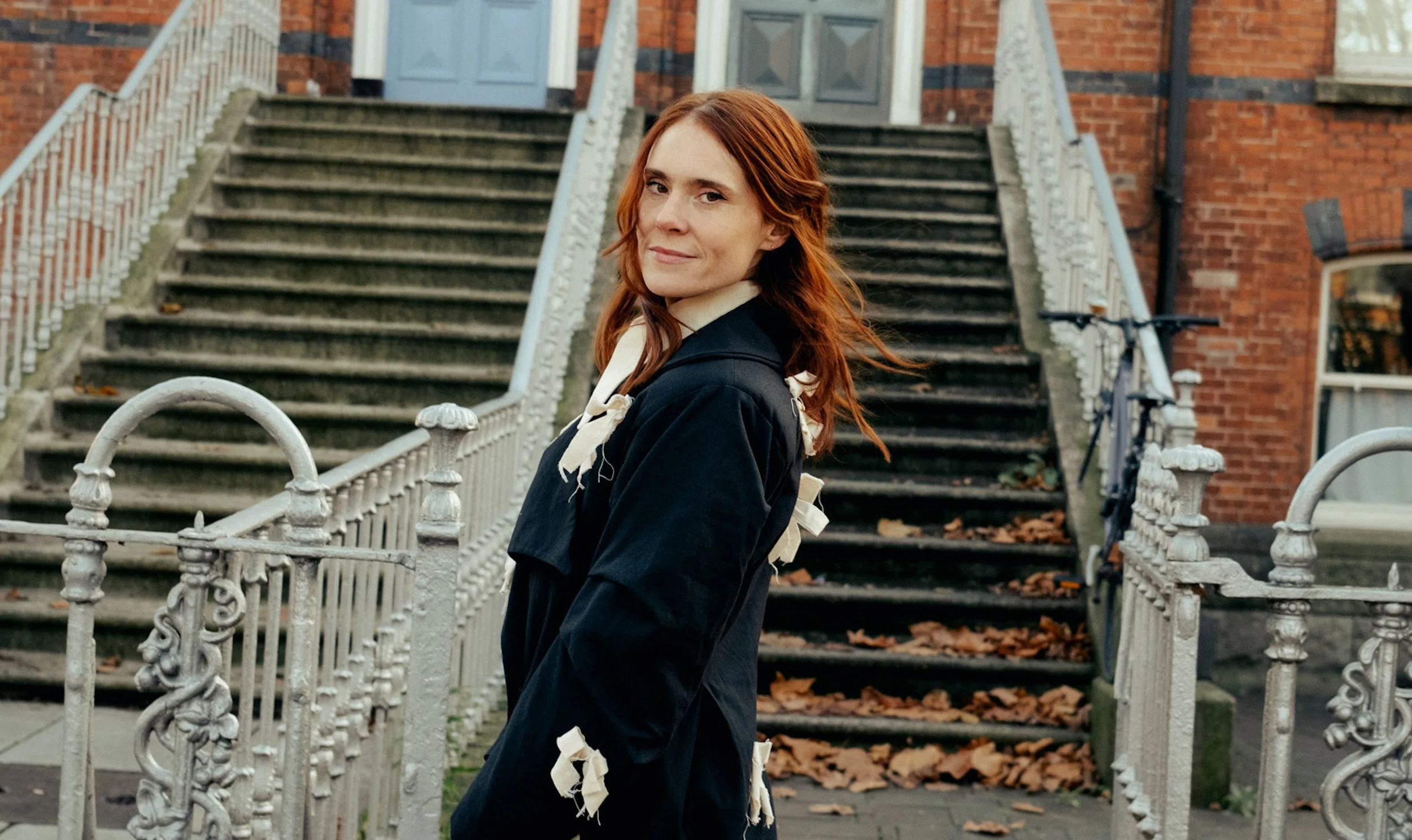 A woman with reddish hair standing in front of a staircase with a white ornate metal railing in an urban setting, with brick buildings and a bicycle in the background.