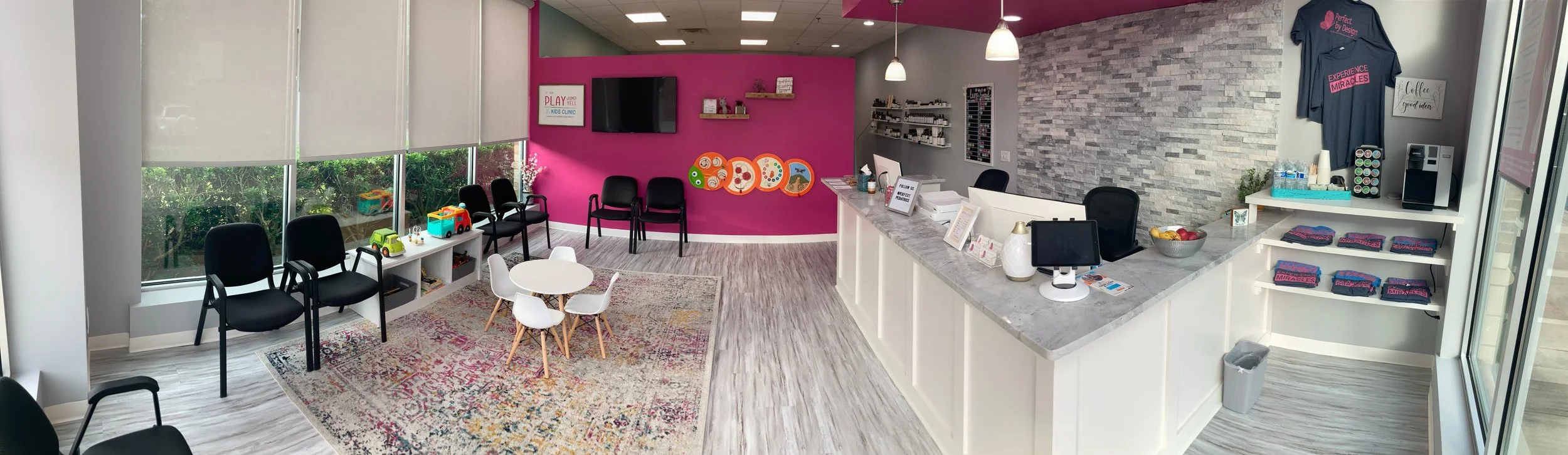 Interior of a modern wellness or pediatric clinic with seating area, colorful wall and a reception desk. Features include black and white chairs, a small round table with children’s toys, a pink accent wall with decorative art, a flat-screen TV, and a reception counter with informational brochures and a snack station.