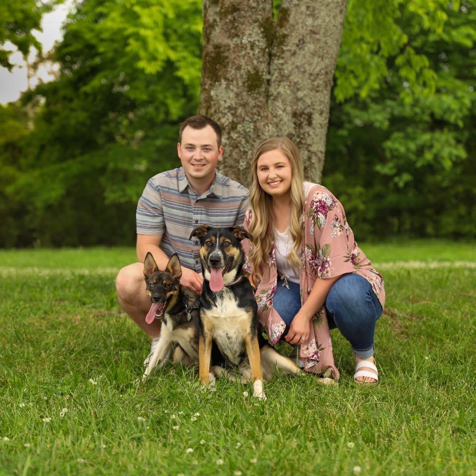 A young man and woman sitting on grass with two dogs in front of a large tree in a park. The man is wearing a striped shirt, and the woman is wearing a floral kimono over a white top and jeans. Both are smiling.