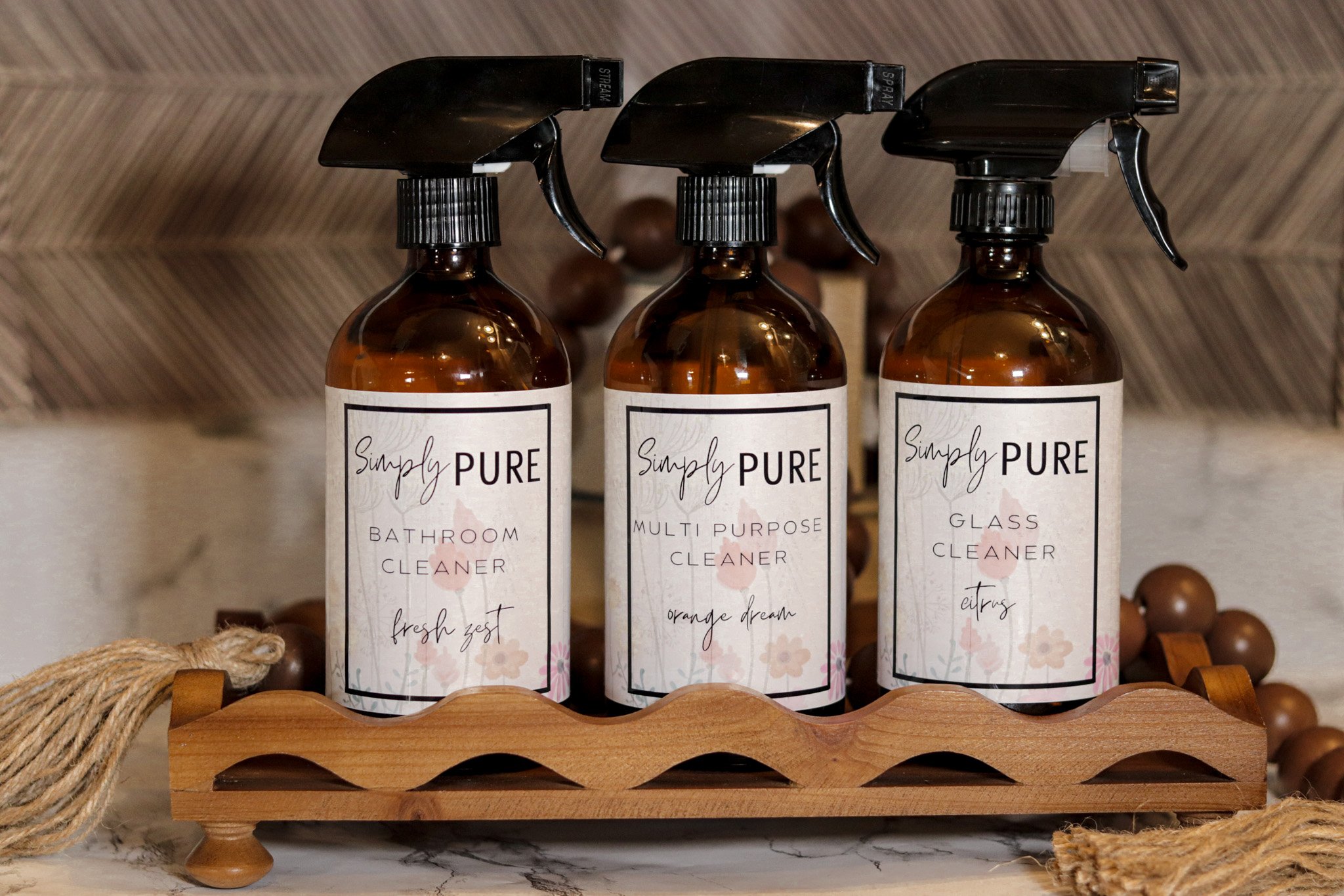 Three amber spray bottles labeled 'Simply Pure' with different cleaning purposes, placed on a wooden tray on a marble surface, against a herringbone-patterned background.