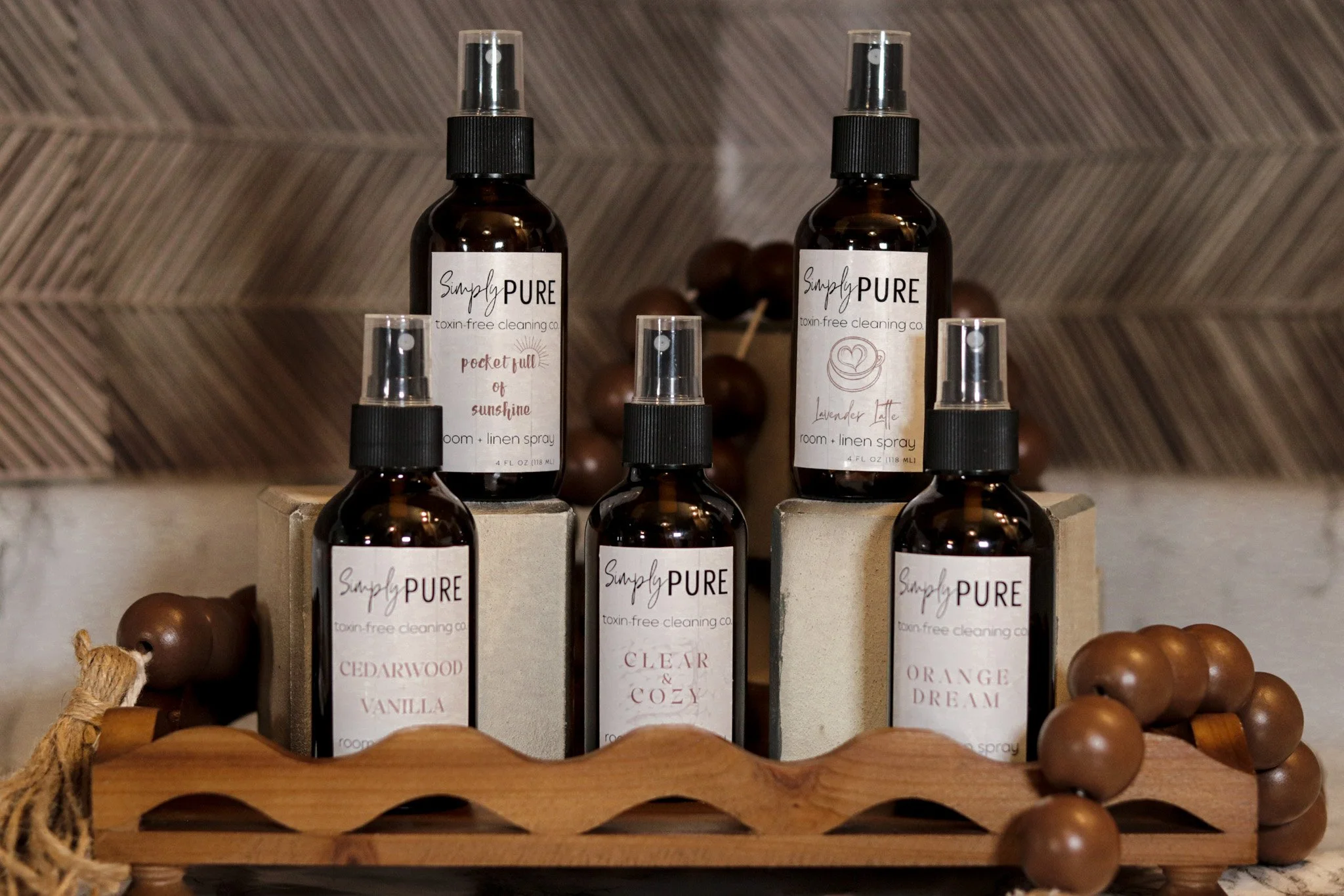 A display of five amber bottles of Simply Pure linen spray with spray nozzles, labeled with scents 'Pocket Full of Sunshine,' 'Lavender Latte,' 'Cedarwood & Vanilla,' 'Clear & Cozy,' and 'Orange Dream.' Behind the bottles are dark brown decorative be