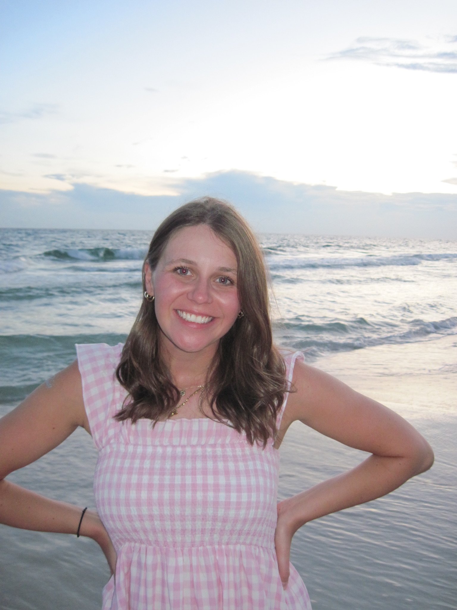 A young woman with long brown hair, smiling, wearing a pink and white checkered dress, standing on a beach near the ocean during sunset.