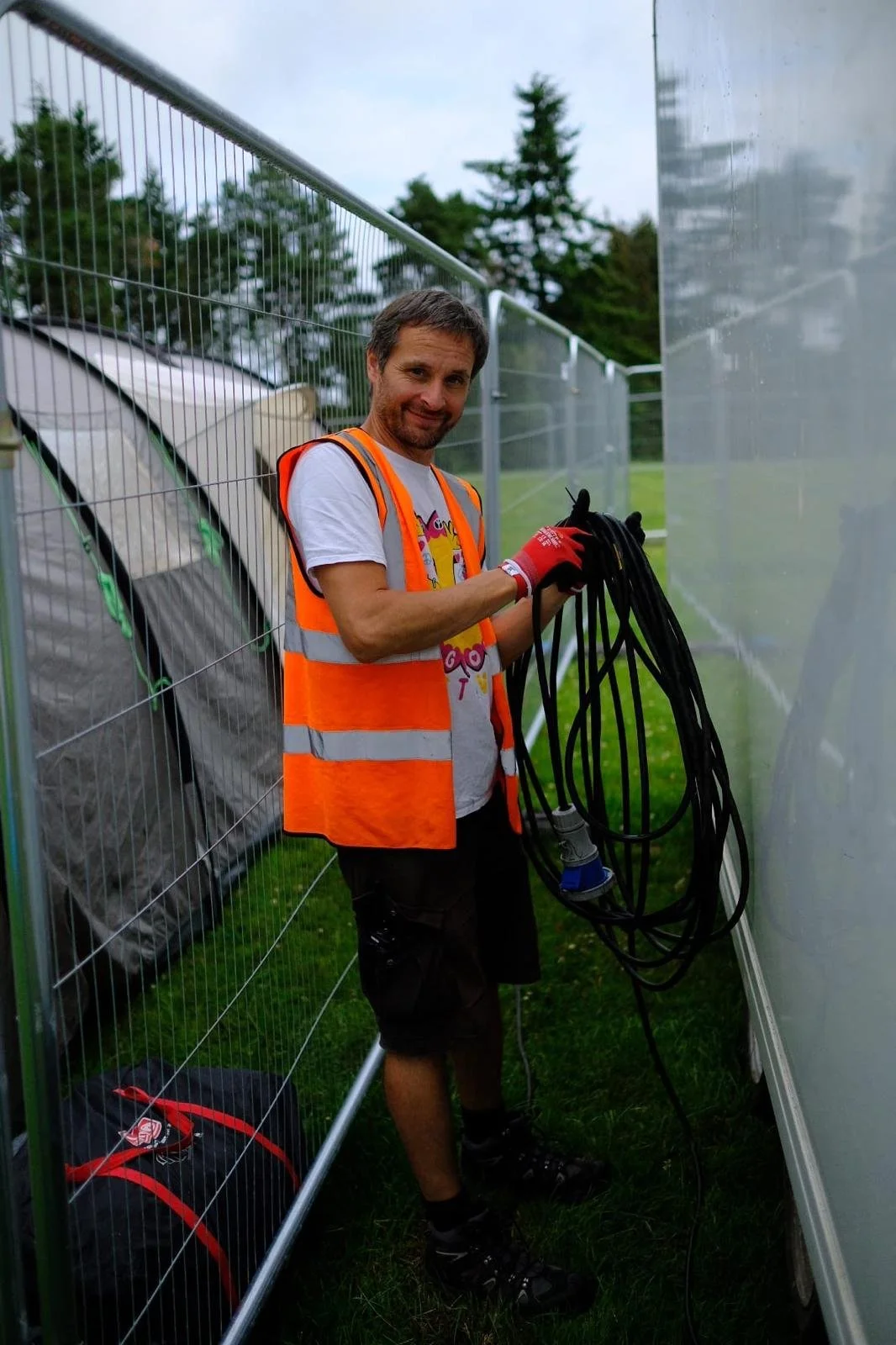 Stu Housley Director & Co Founder of Big Love Festival wearing a gray T-shirt with colorful graphics, an orange safety vest, red gloves, and black shorts stands outside between a wire fence and a reflective wall, holding black electrical cords.
