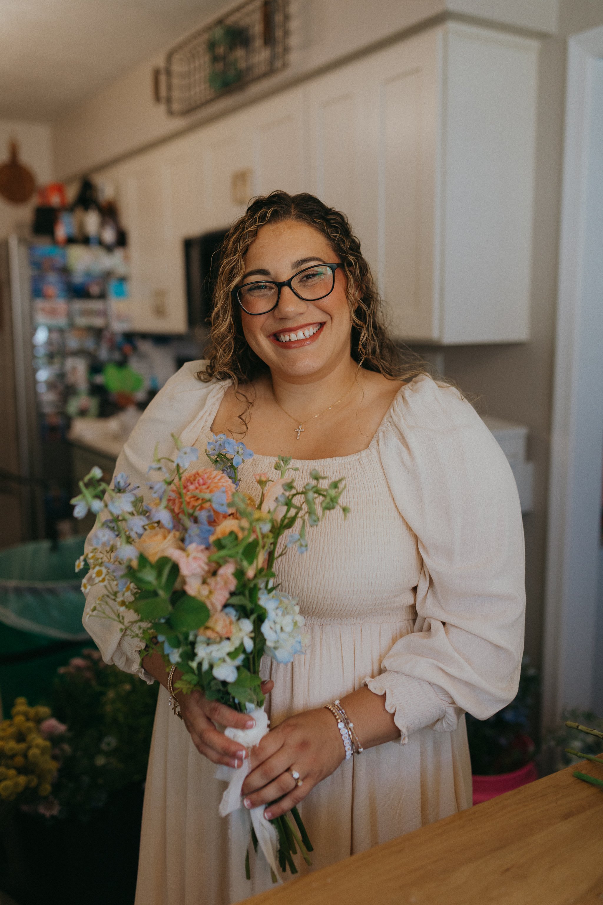 A woman with curly hair, glasses, and a big smile holding a bouquet of colorful flowers inside a house.