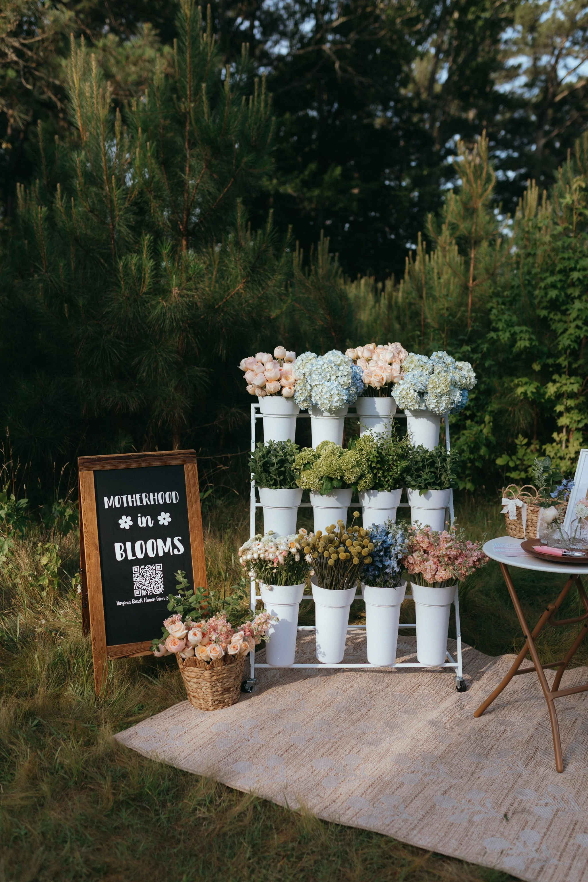 A display of white flower pots filled with pink, white, and green flowers, arranged on a white metal stand on a woven rug outdoors. A chalkboard sign reads "Motherhood in Bloom" with a QR code below, and a wicker basket with pink roses is nearby. Trees and bushes surround the scene.