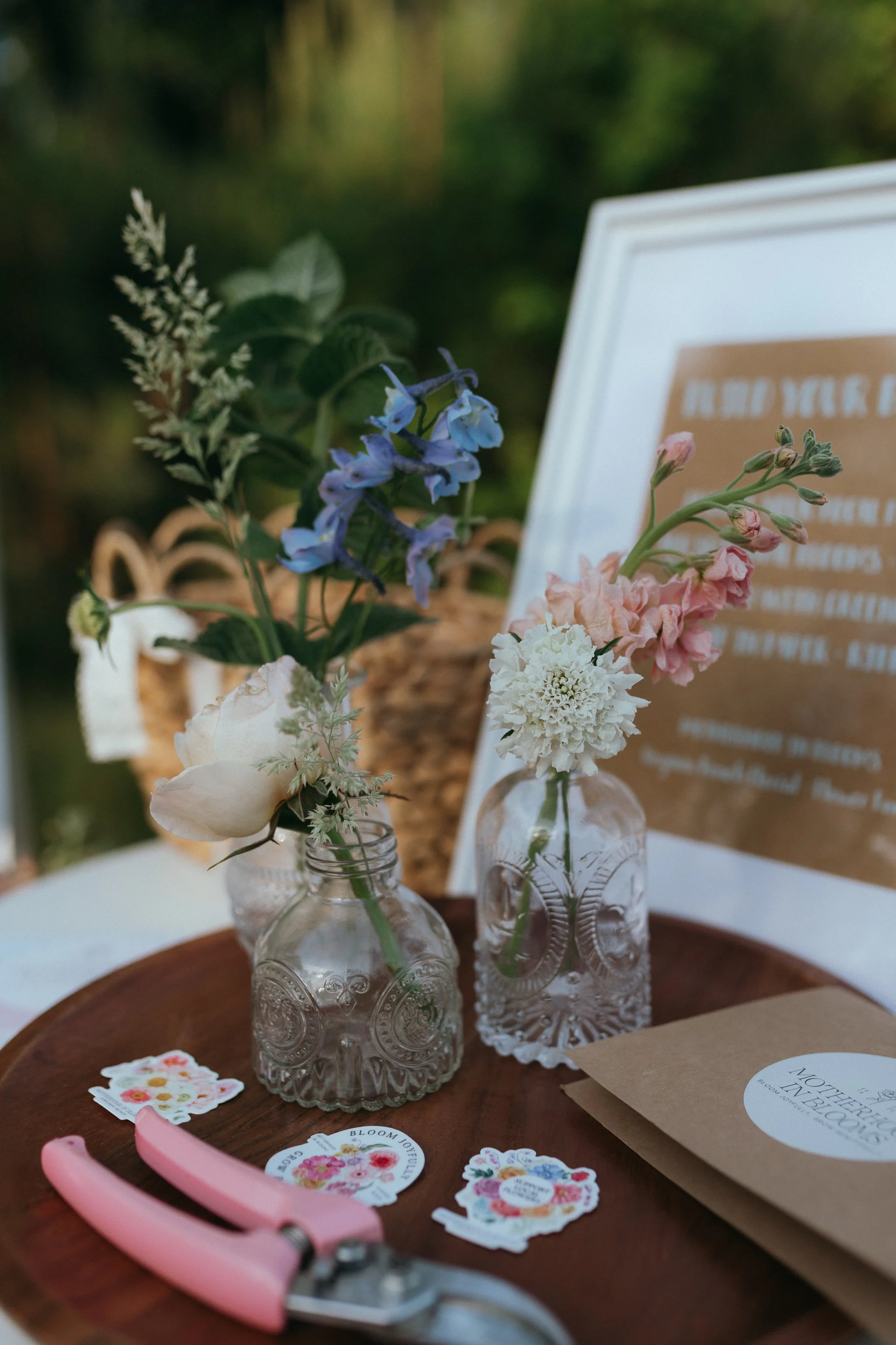 Close-up of a floral arrangement in vintage glass bottles on a wooden tray, with small decorative stickers and a pink miniature handled tool on a table; out of focus background with a framed message.