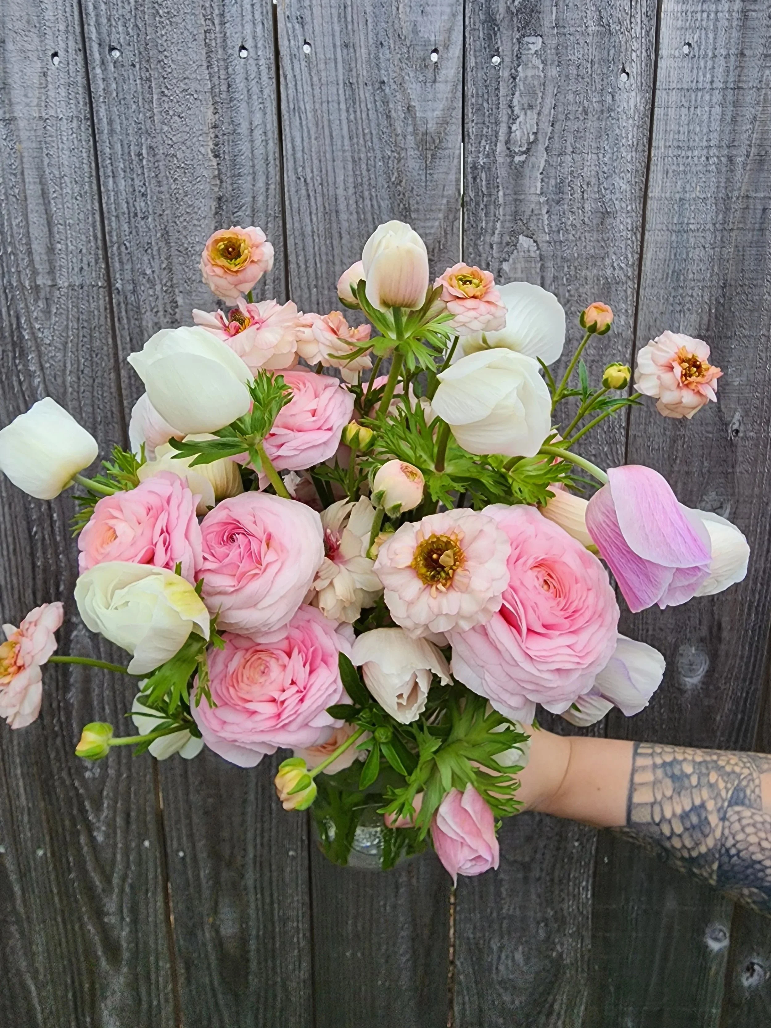 A bouquet of pink and white flowers held by a person with a tattoo on their arm, against a rustic wooden fence background.
