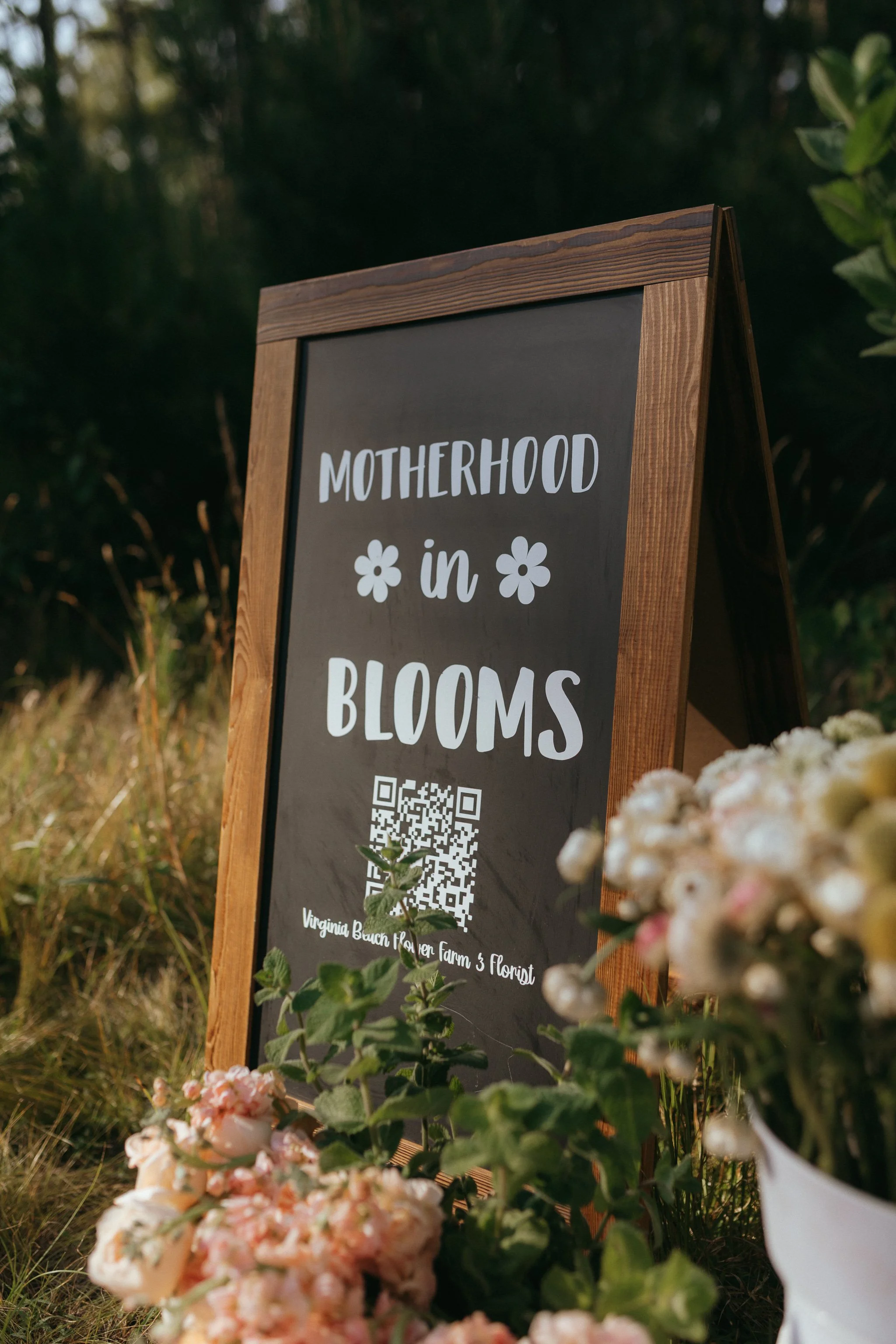 Wooden framed chalkboard sign reading 'Motherhood in Blooms' with a QR code, surrounded by pink and white flowers outdoors with greenery in the background.