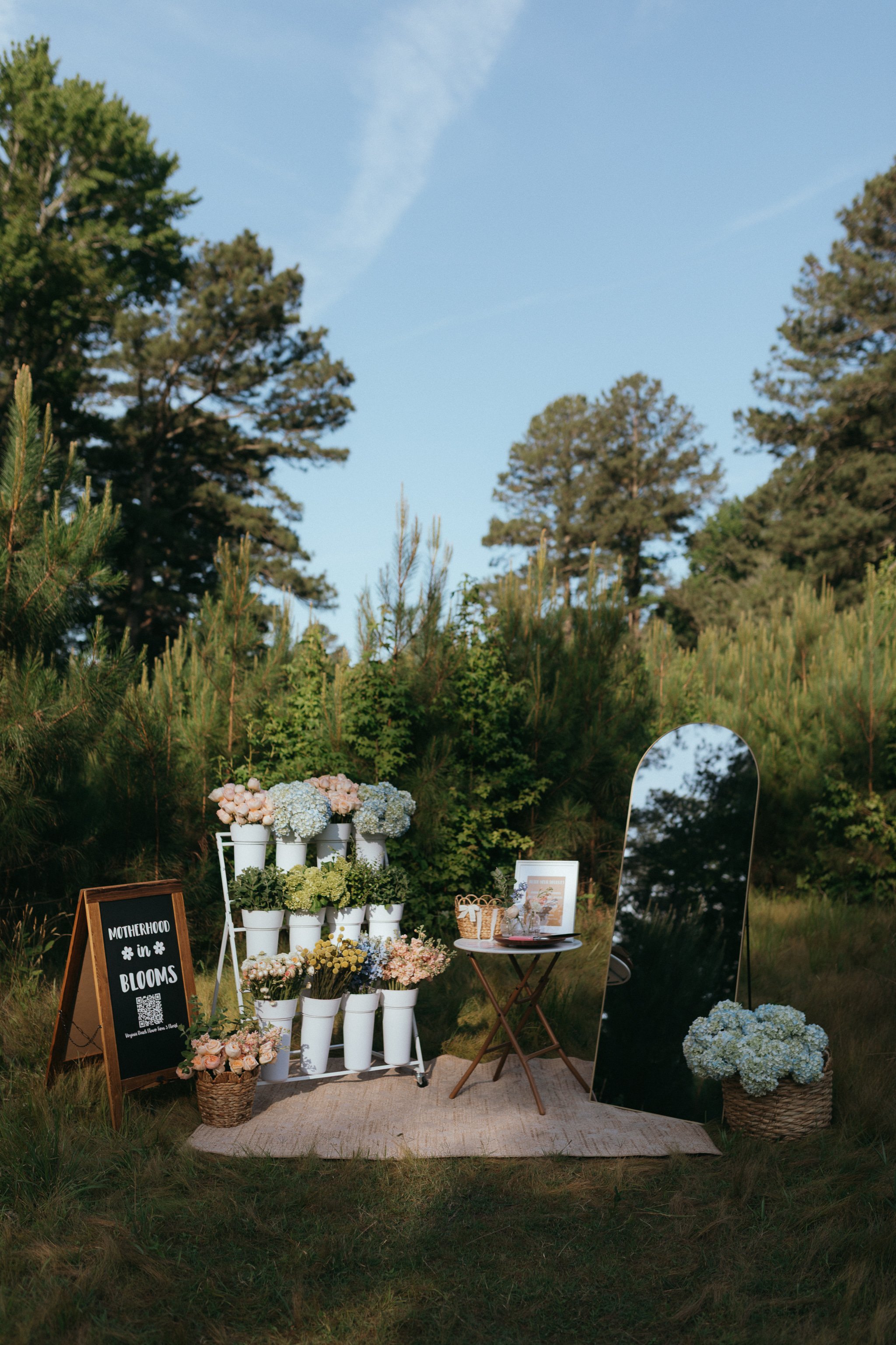 Outdoor floral display with white flower arrangements in vases, a chalkboard sign reading 'Motherhood in Blooms,' and a reflective mirror set in a natural setting with trees and grass.