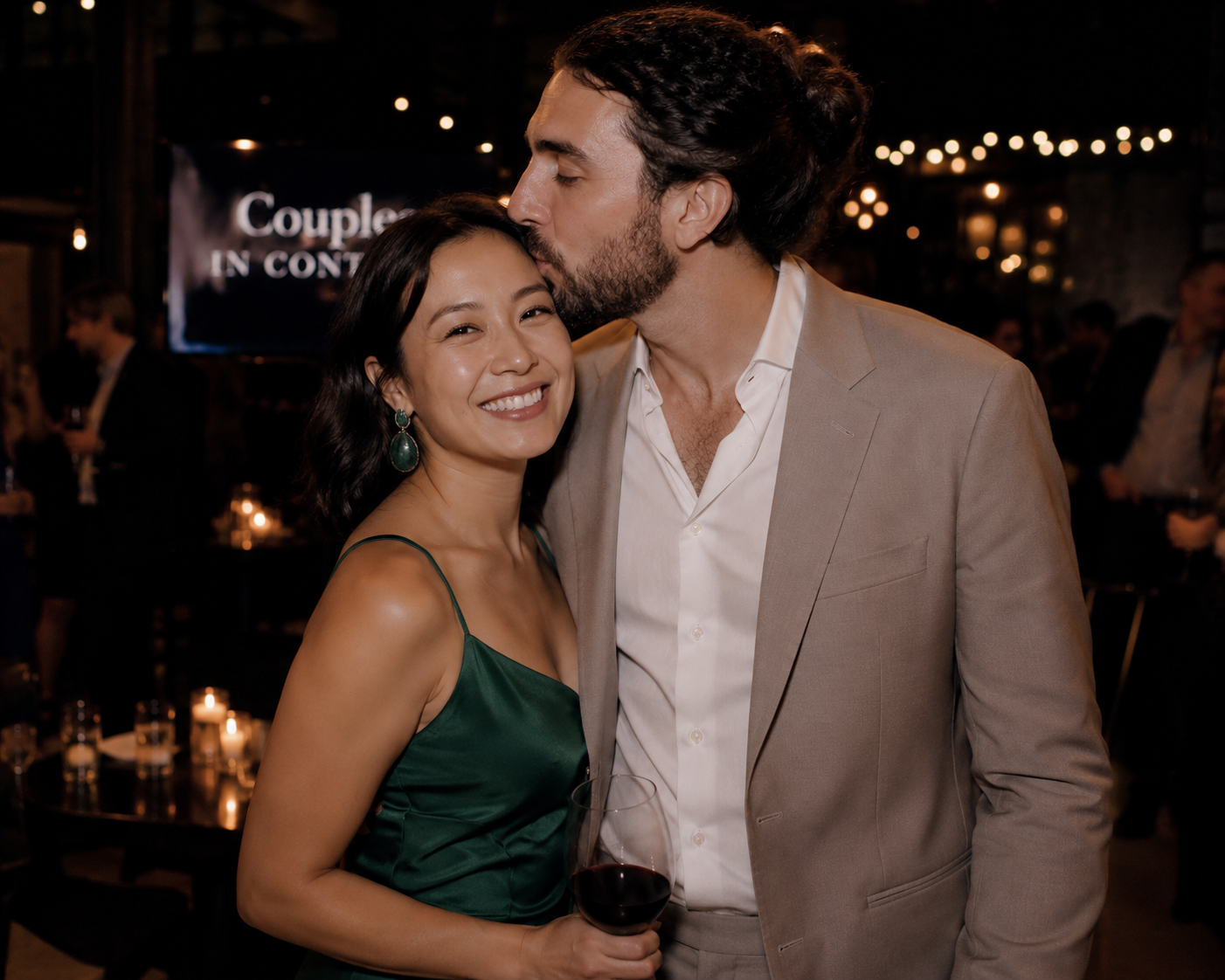 A couple at a nighttime event, with the man kissing the woman's forehead. The woman is smiling, wearing a green satin dress and large earrings, holding a glass of red wine. The man is in a beige suit with a white shirt. The background is dimly lit with candles and string lights.