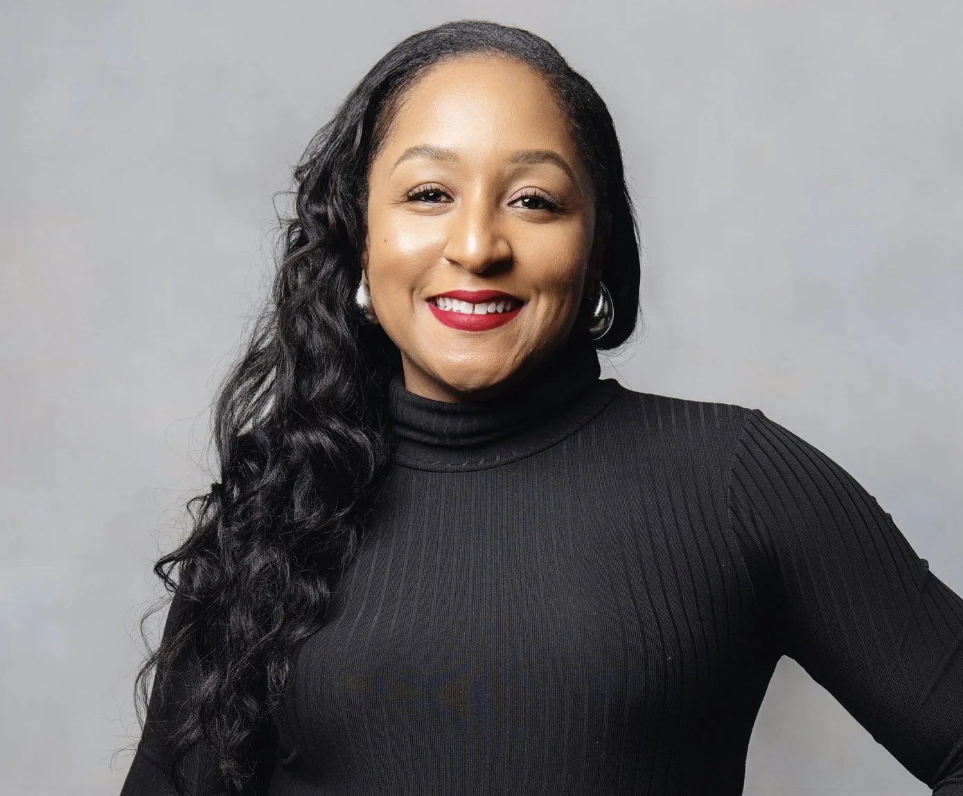 A smiling woman with long curly black hair, wearing a black turtleneck, red lipstick, and silver hoop earrings, against a plain light gray background.