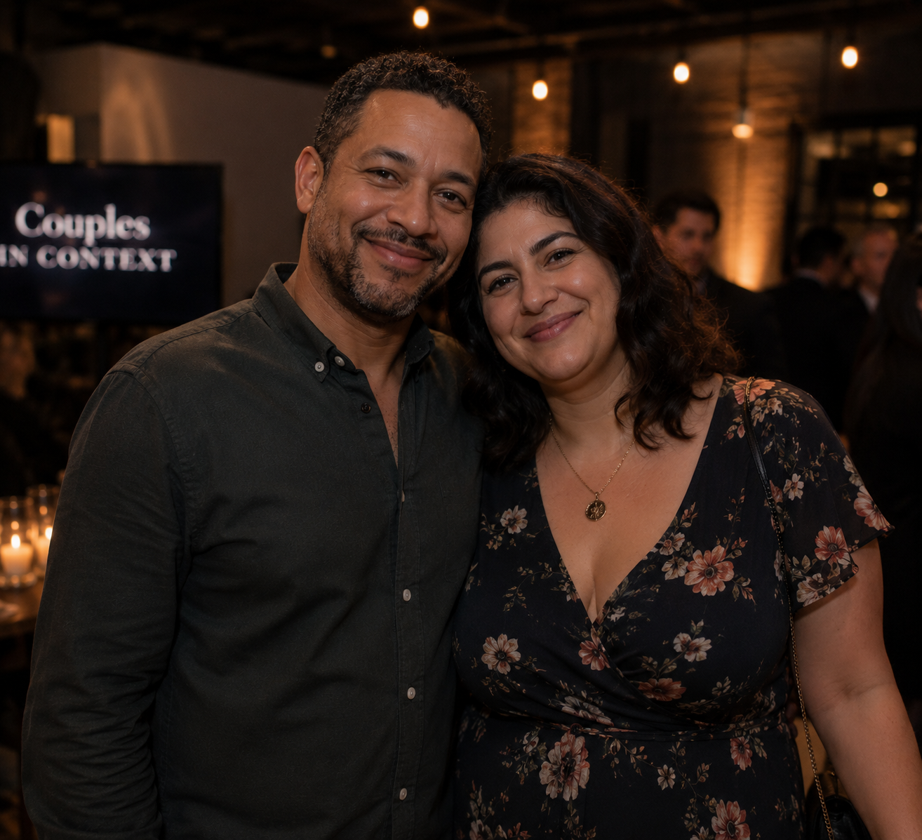 A smiling man and woman posing together at a social event in a dimly-lit venue with Edison bulb lighting, with a screen in the background displaying the words 'Couples in Context.'