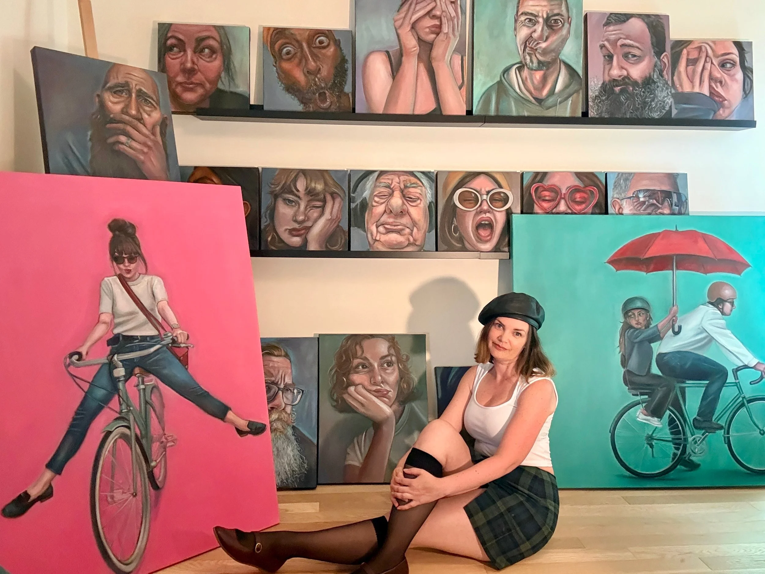 Young woman with long brown hair, wearing a white tank top, plaid skirt, black knee-high socks, and black boots, sitting on the floor with her legs crossed, smiling at the camera in front of colorful artwork of people with expressive faces, some holding bicycles, and others with exaggerated facial expressions.